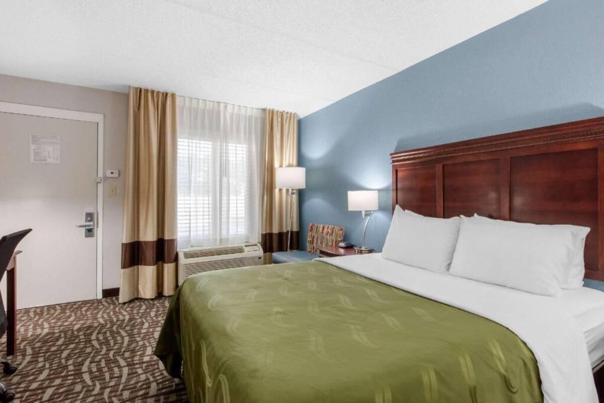 A hotel room with a bed, a desk, a chair, a lamp, and a window with curtains.