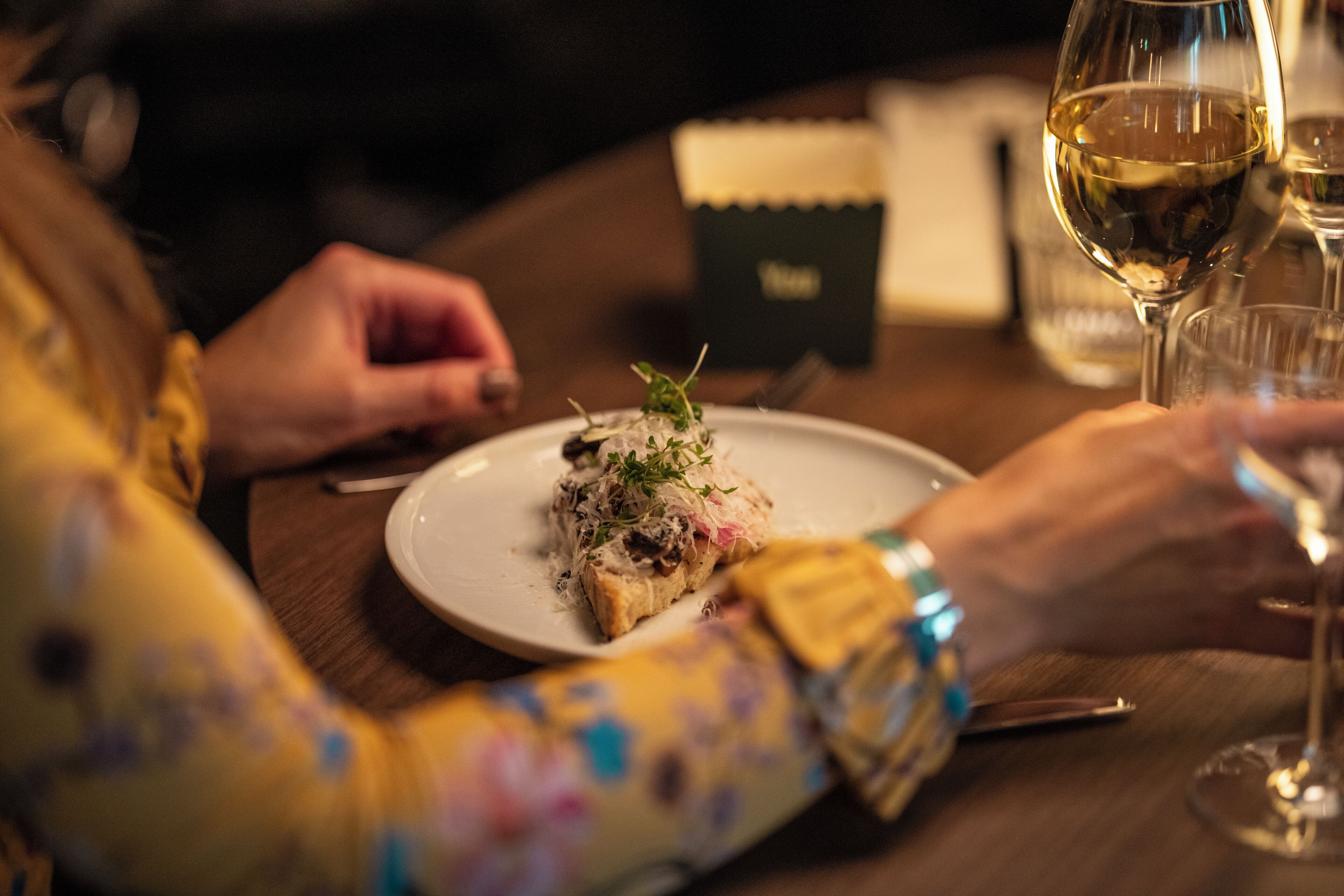 A person dining at a table with a plate of food and a glass of wine.