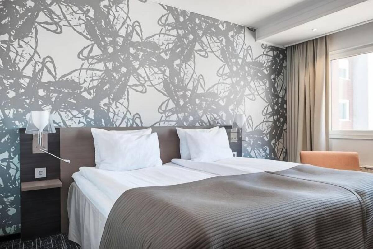 Una habitación de hotel con cama, mesita de noche, lámpara y ventana con cortinas.