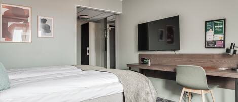 A hotel room with a bed, a desk, a chair, a TV, and a poster on the wall.