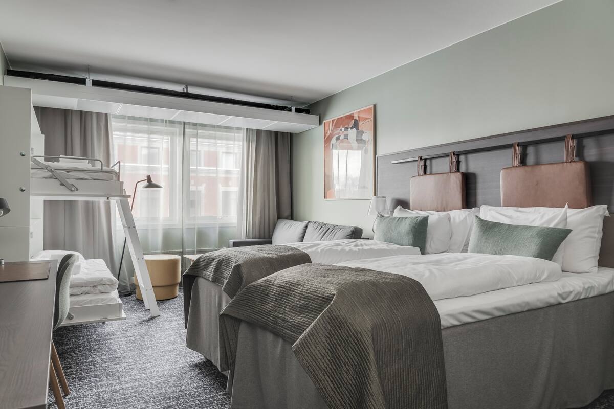 Una habitación de hotel con literas, un escritorio, una silla, una lámpara y un cuadro en la pared.