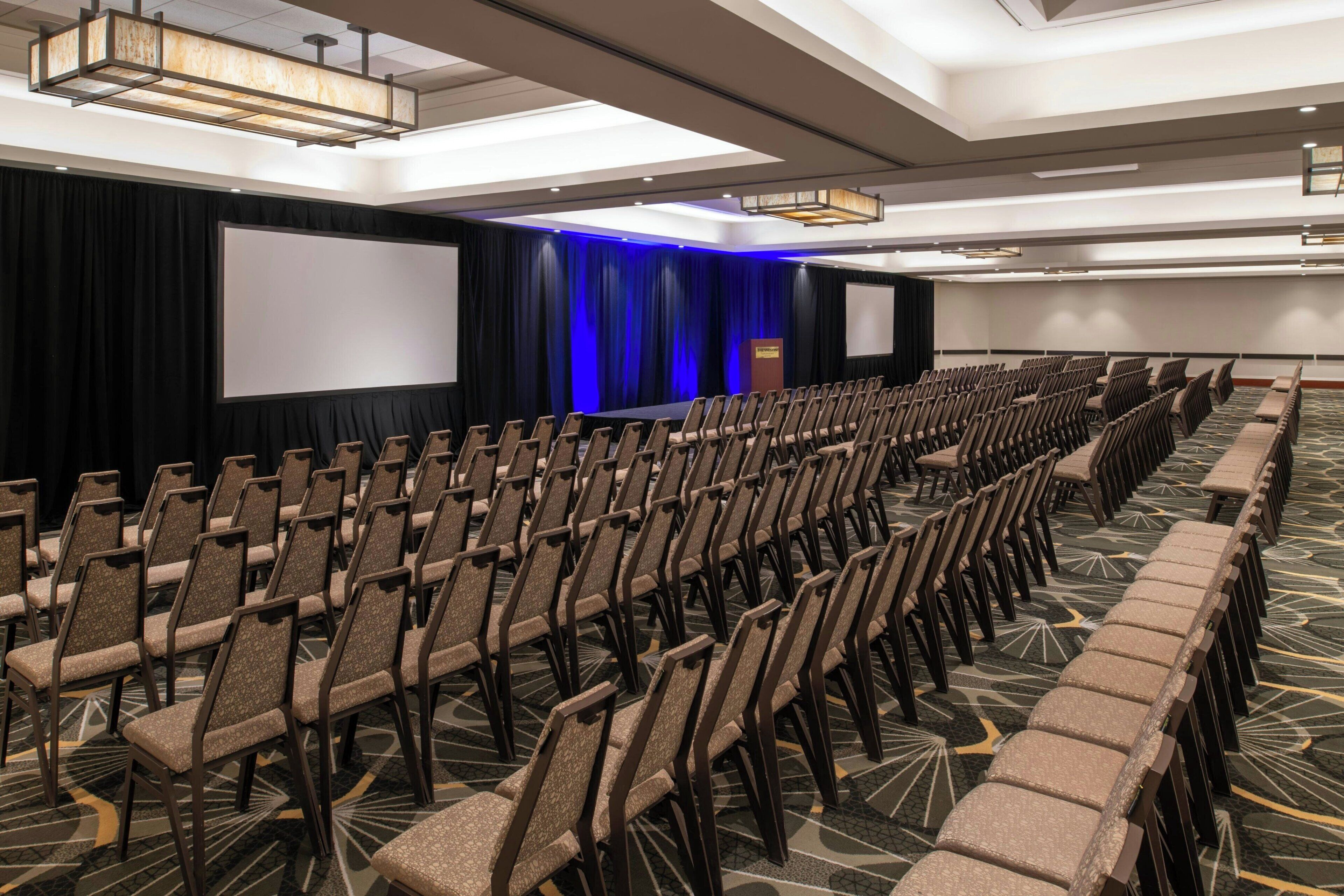 A conference room with rows of chairs, a stage with a blue curtain backdrop, and a large screen.