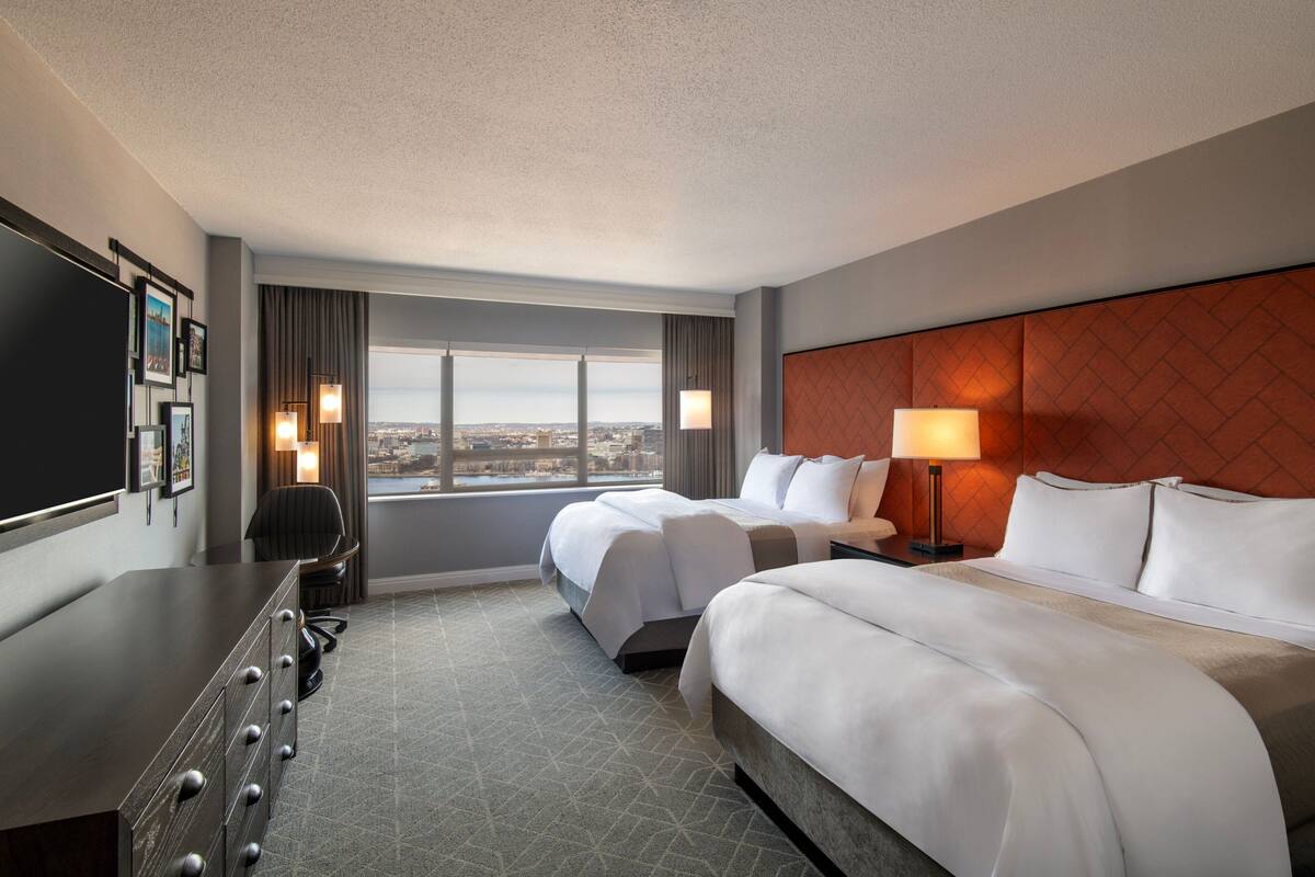 Habitación de hotel con dos camas, un escritorio, televisor y vista a la ciudad.