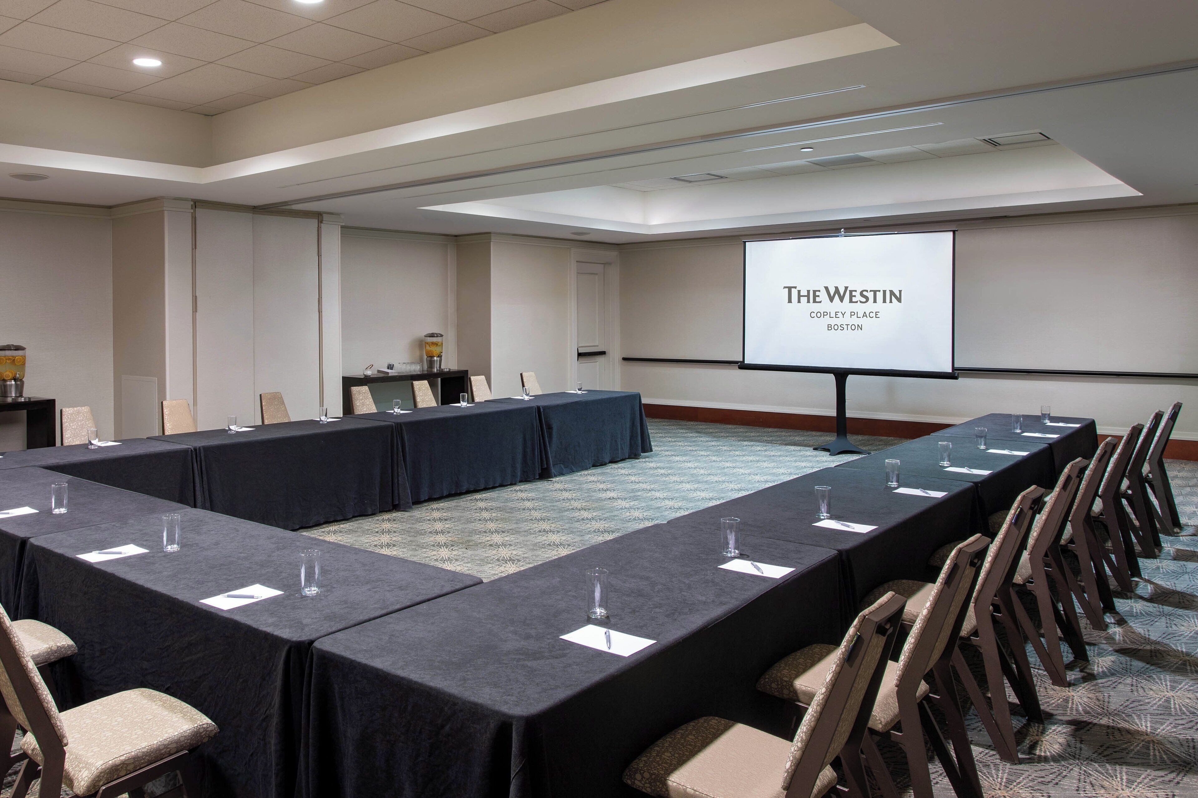 A conference room with a long table, chairs, a projector screen displaying "The Westin Copley Place Boston", and a small table with a coffee machine.