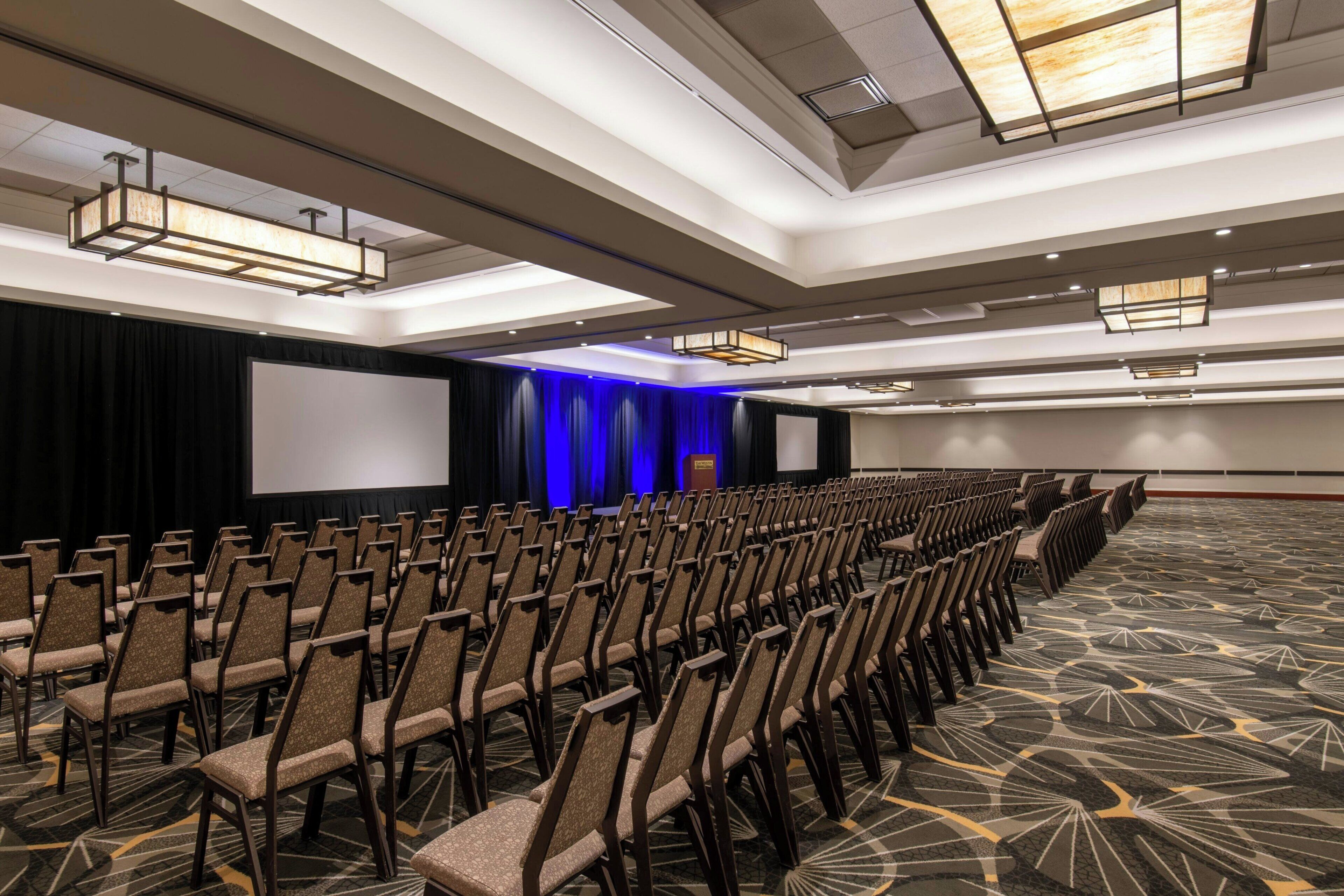 A conference room with rows of chairs, a stage with curtains, a projector screen, and a patterned carpet.