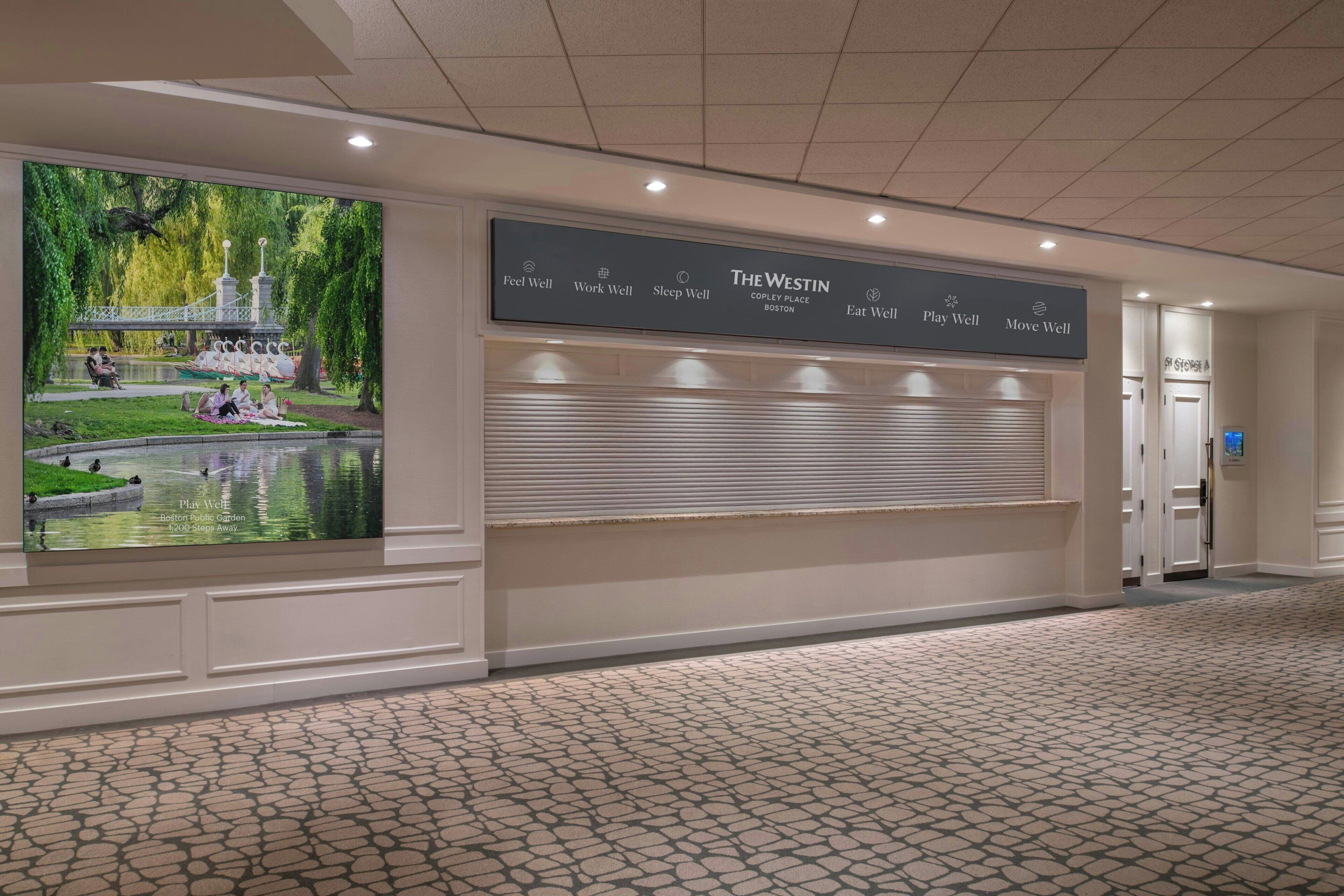 A hotel corridor with a large mural of a park, a reception desk, and a sign with wellness-related phrases.