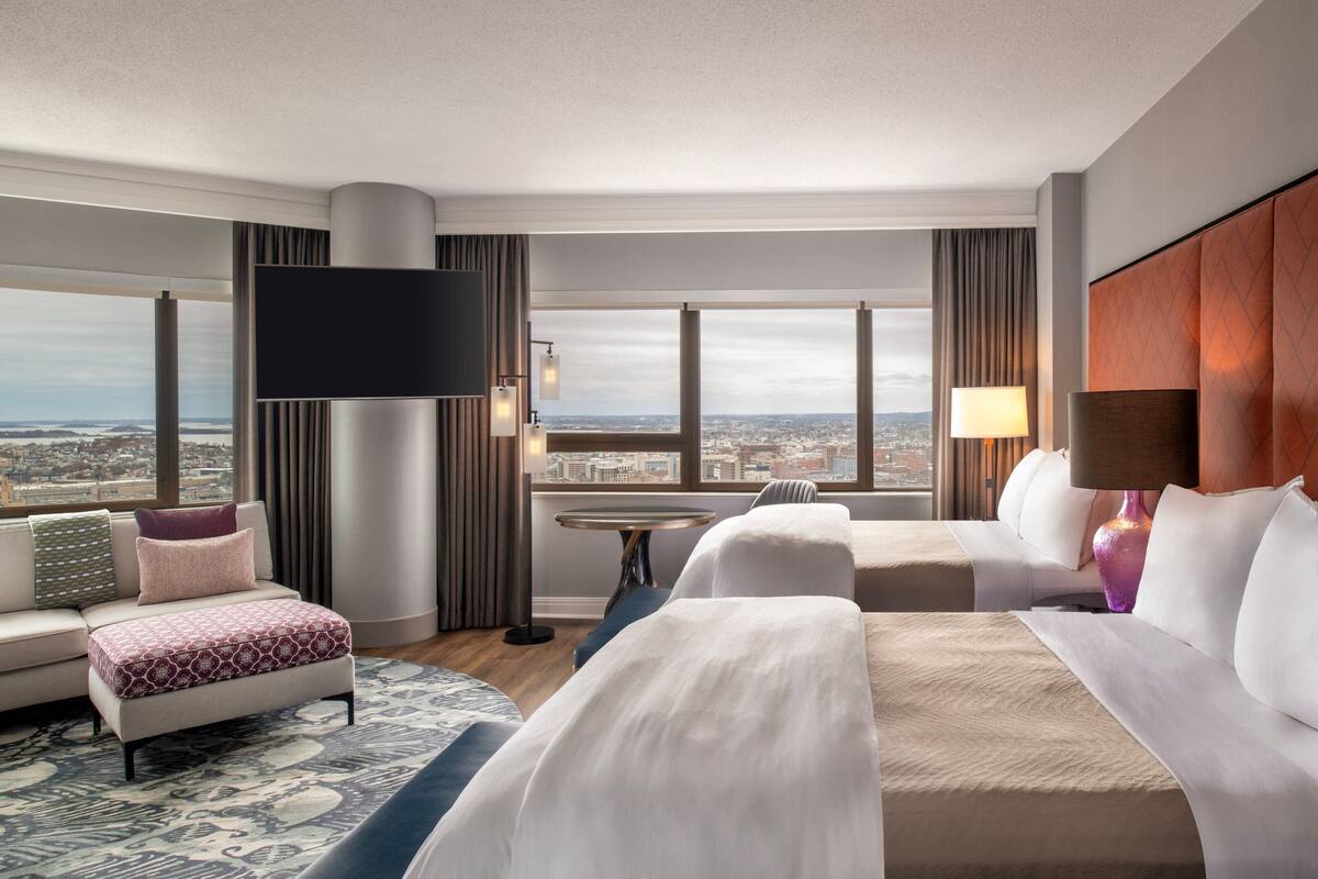 Habitación de hotel con una cama grande, un sofá, un escritorio y un televisor. Amplias ventanas con vistas a la ciudad.