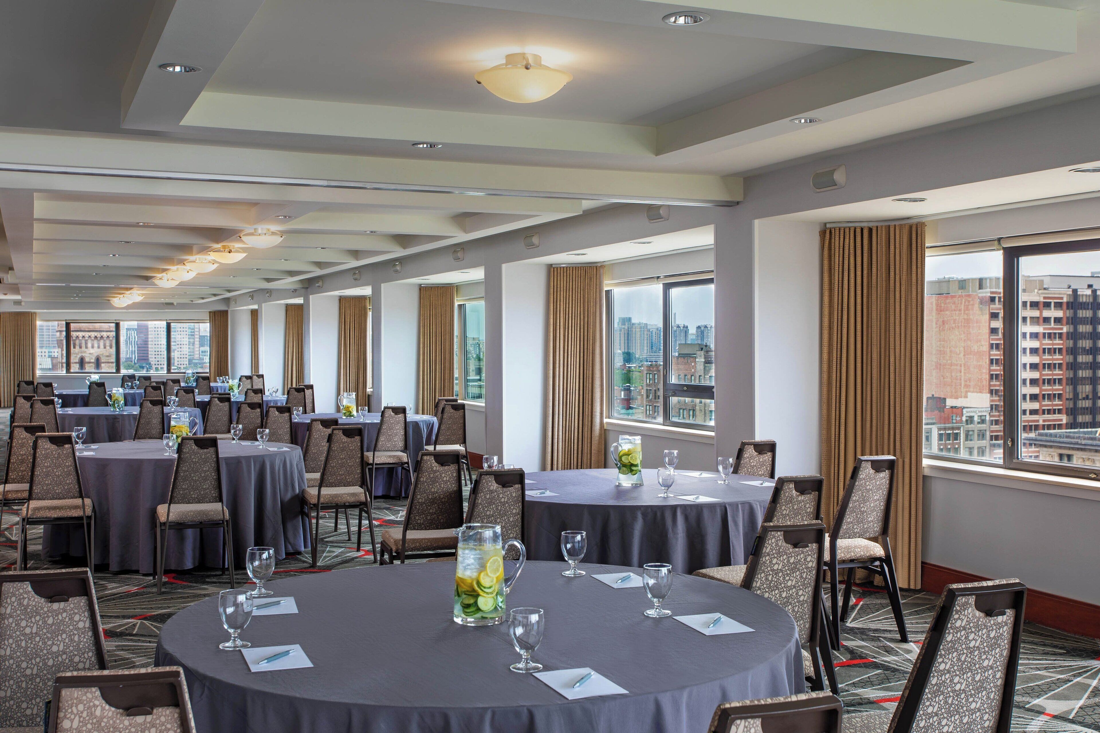 A conference room with multiple tables and chairs, large windows with curtains, and a city view.