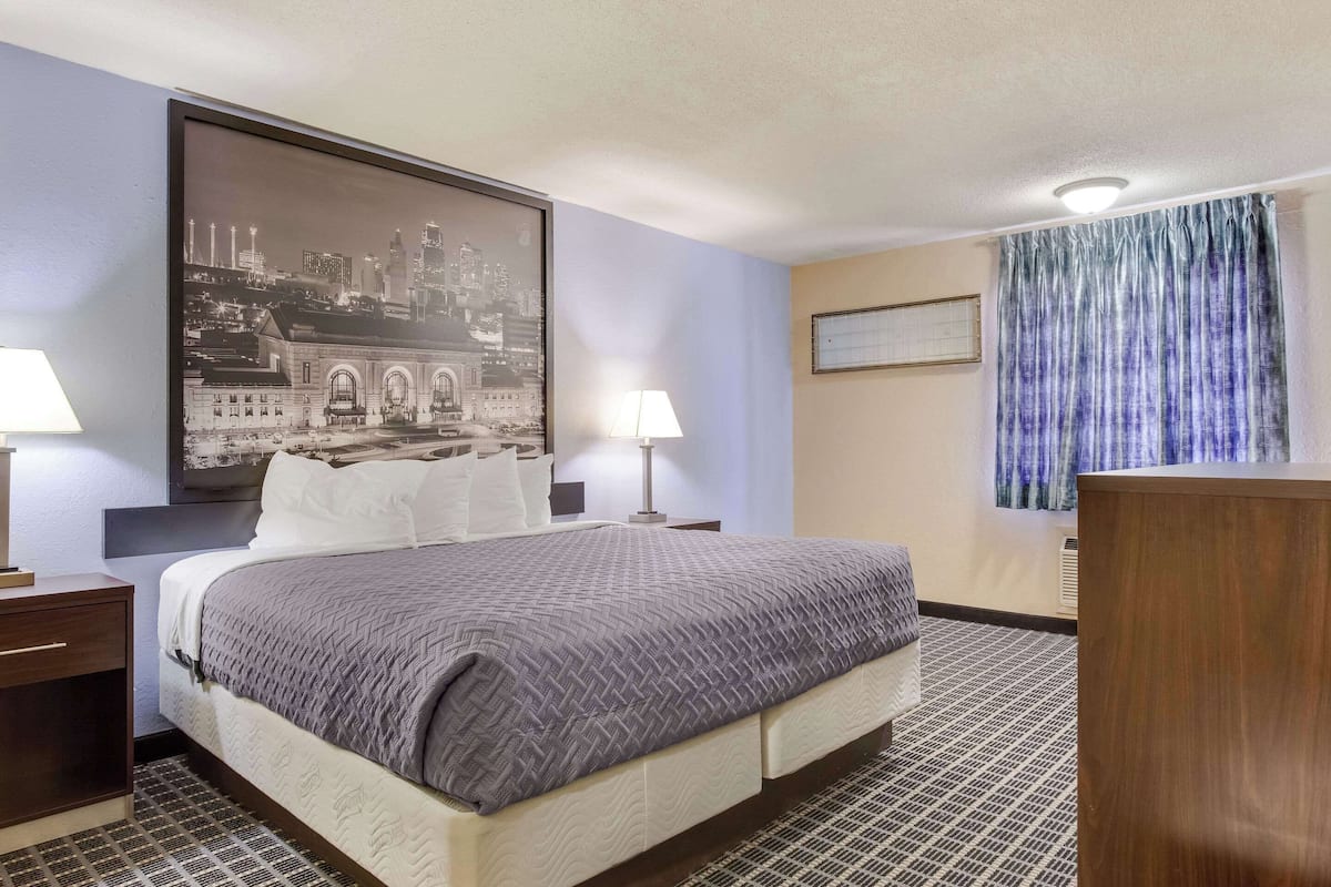 Ein Hotelzimmer mit einem großen Bett, zwei Nachttischen, einem kleinen Tischchen, einem Stadtbild in einem Rahmen, einem Fenster mit blauen Vorhängen und einem Holztisch.