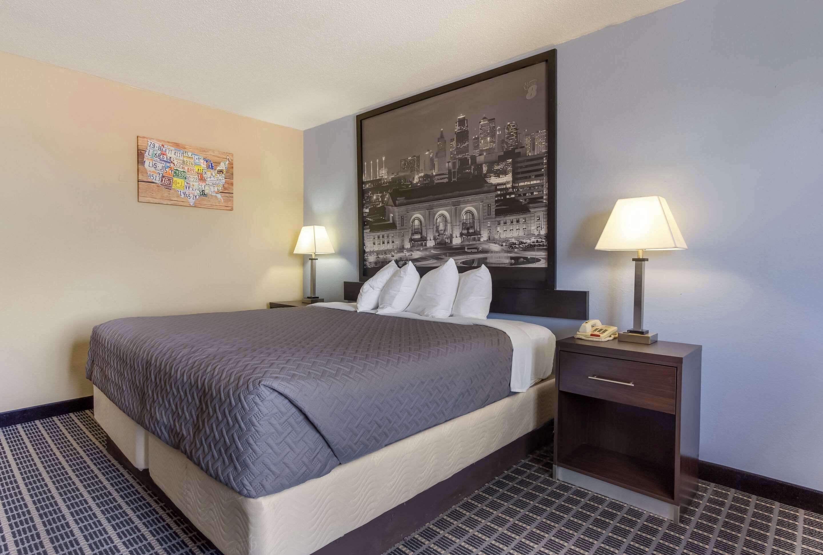 A hotel room with a large bed, two bedside tables with lamps, a nightstand with a phone, and a cityscape artwork on the wall.
