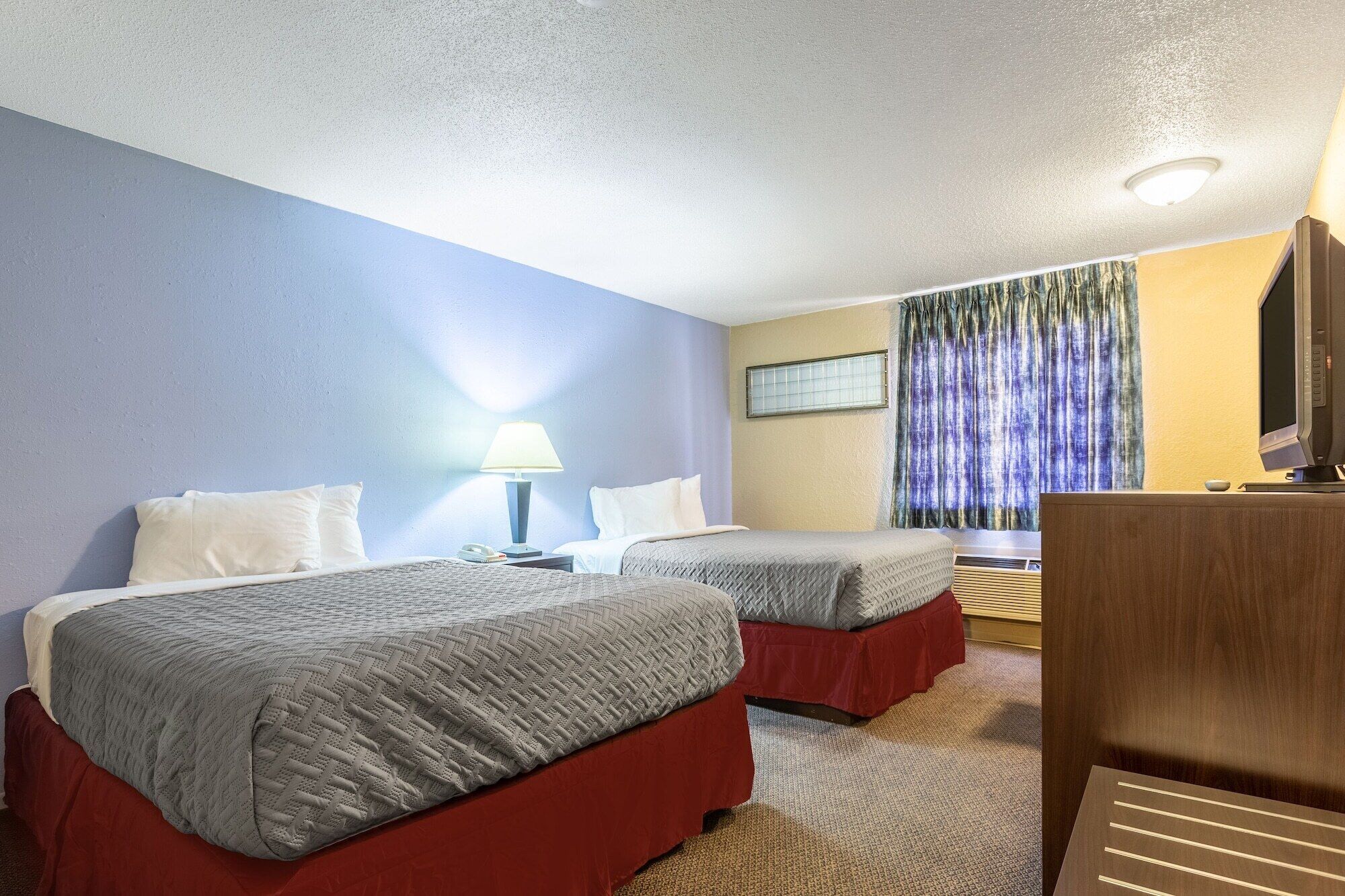 Habitación de hotel con dos camas, un escritorio, una lámpara, un televisor y una ventana con cortinas.
