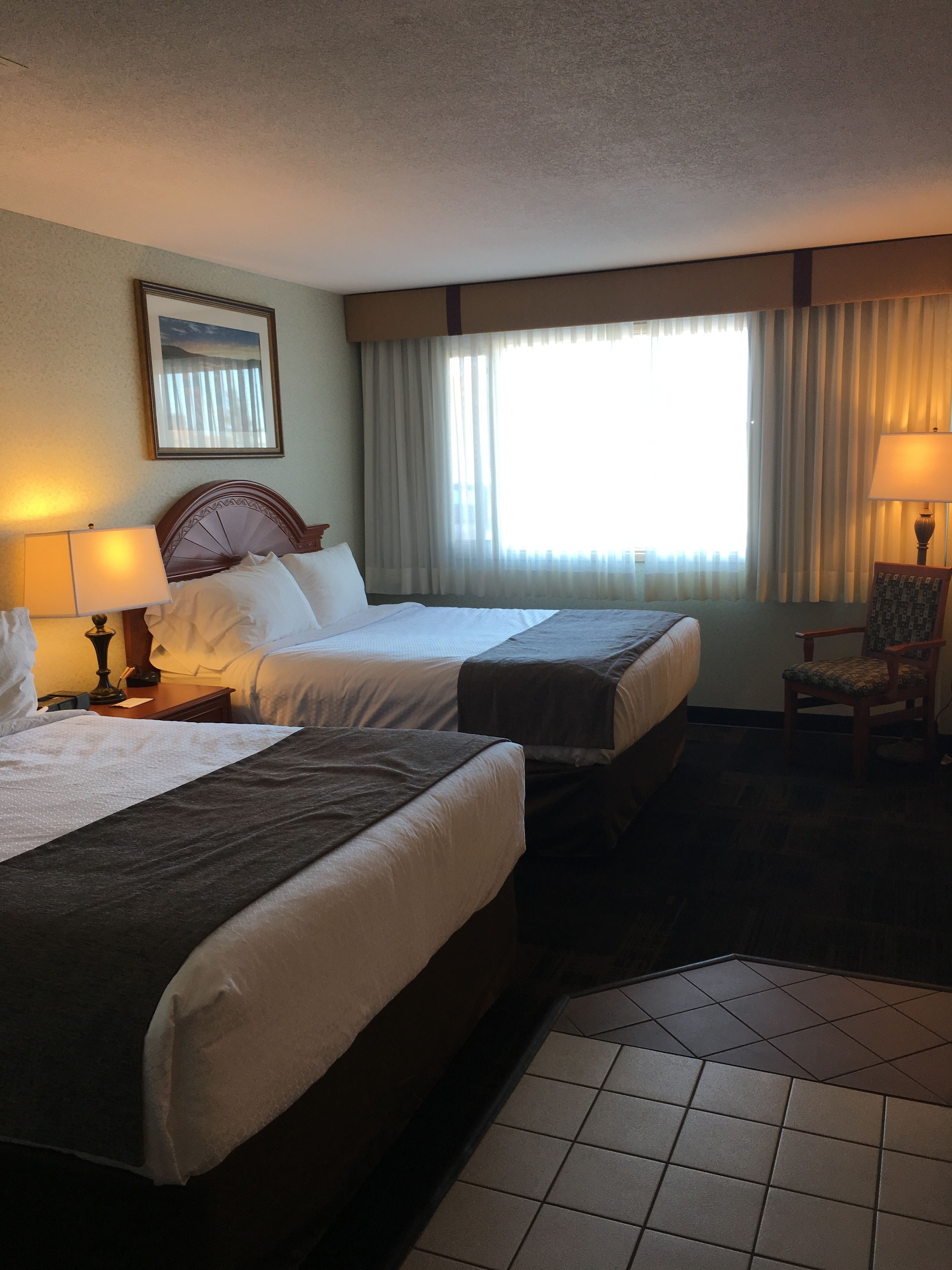 A hotel room with two beds, a chair, a lamp, a painting on the wall, and a window with curtains.