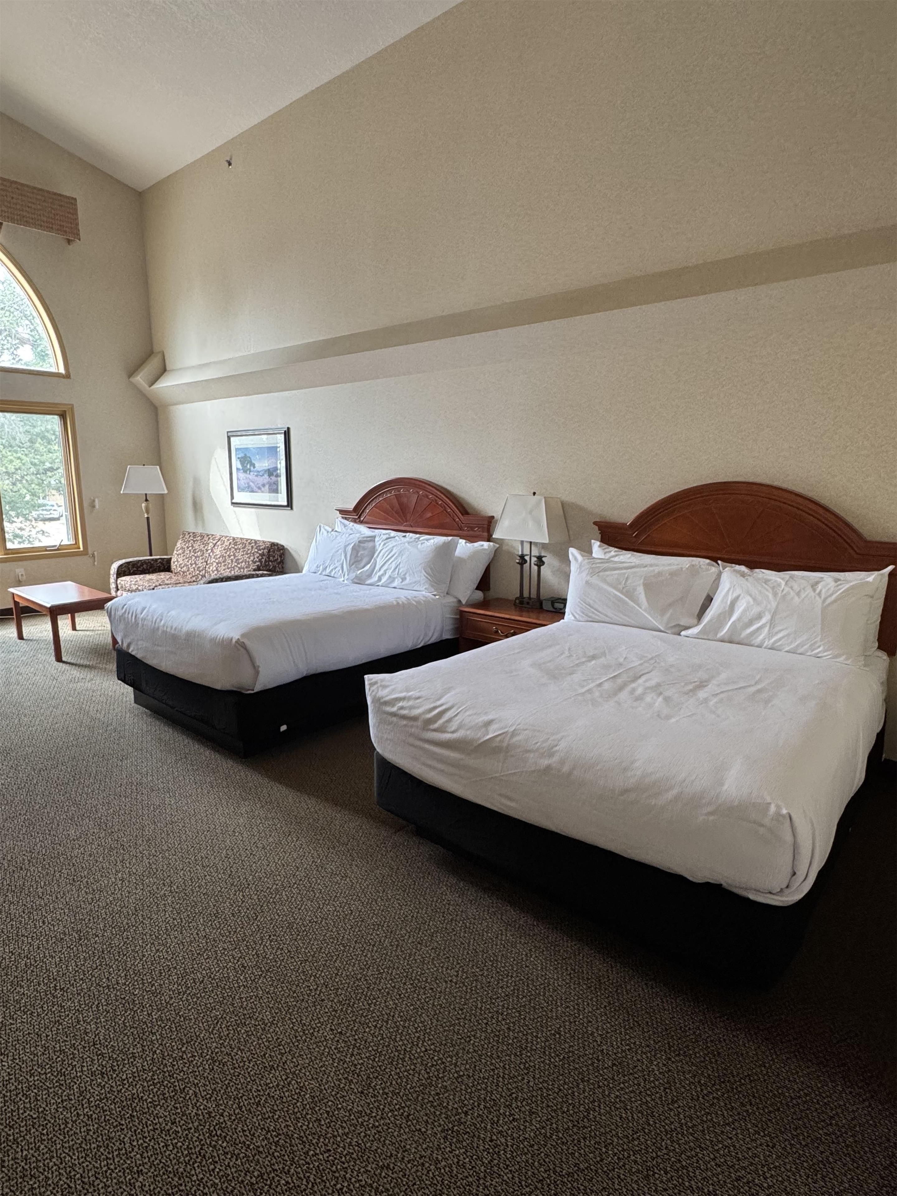 Habitación de hotel con dos camas, un ventanal grande, un cuadro en la pared y una mesita con una lámpara.