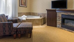 A hotel room with a large bathtub, a flat-screen TV mounted above a fireplace, and patterned armchairs. - Flamingo Hotel (Medicine Hat)