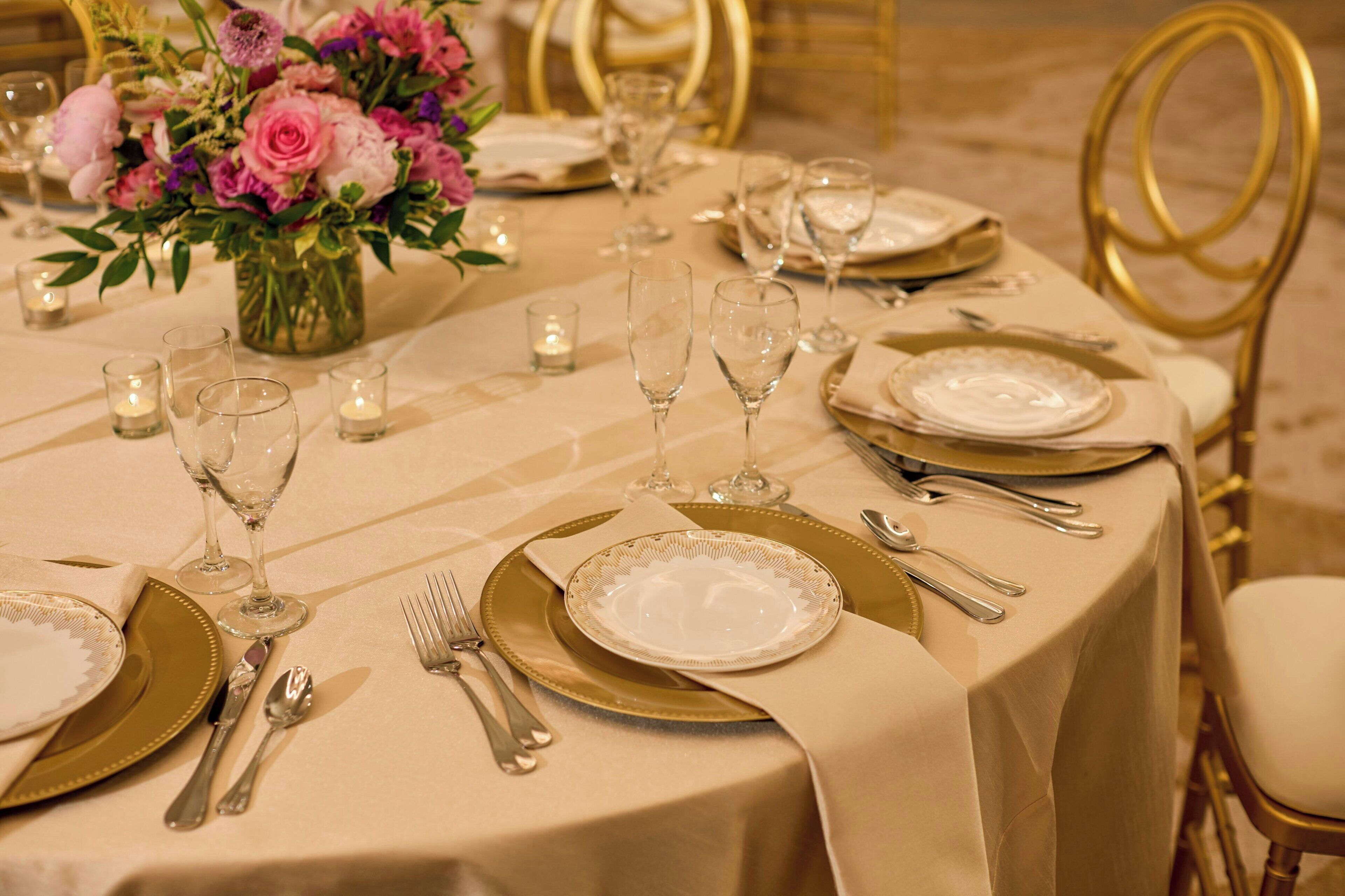 Una mesa dispuesta para una cena formal, con vajilla dorada y blanca, un jarrón de flores y velas.