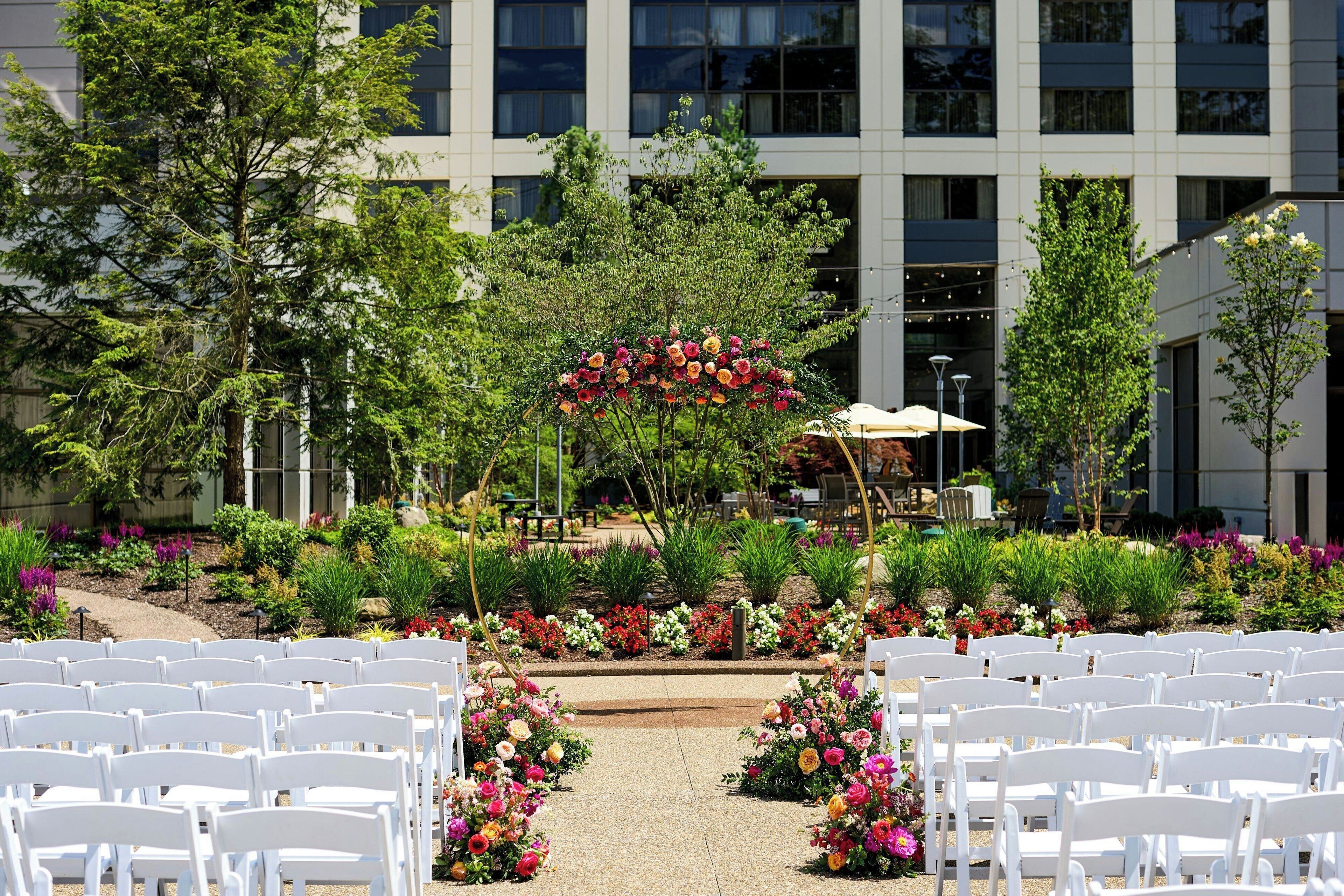 An outdoor ceremony area with white chairs arranged in rows, surrounded by a garden with various plants and flowers.