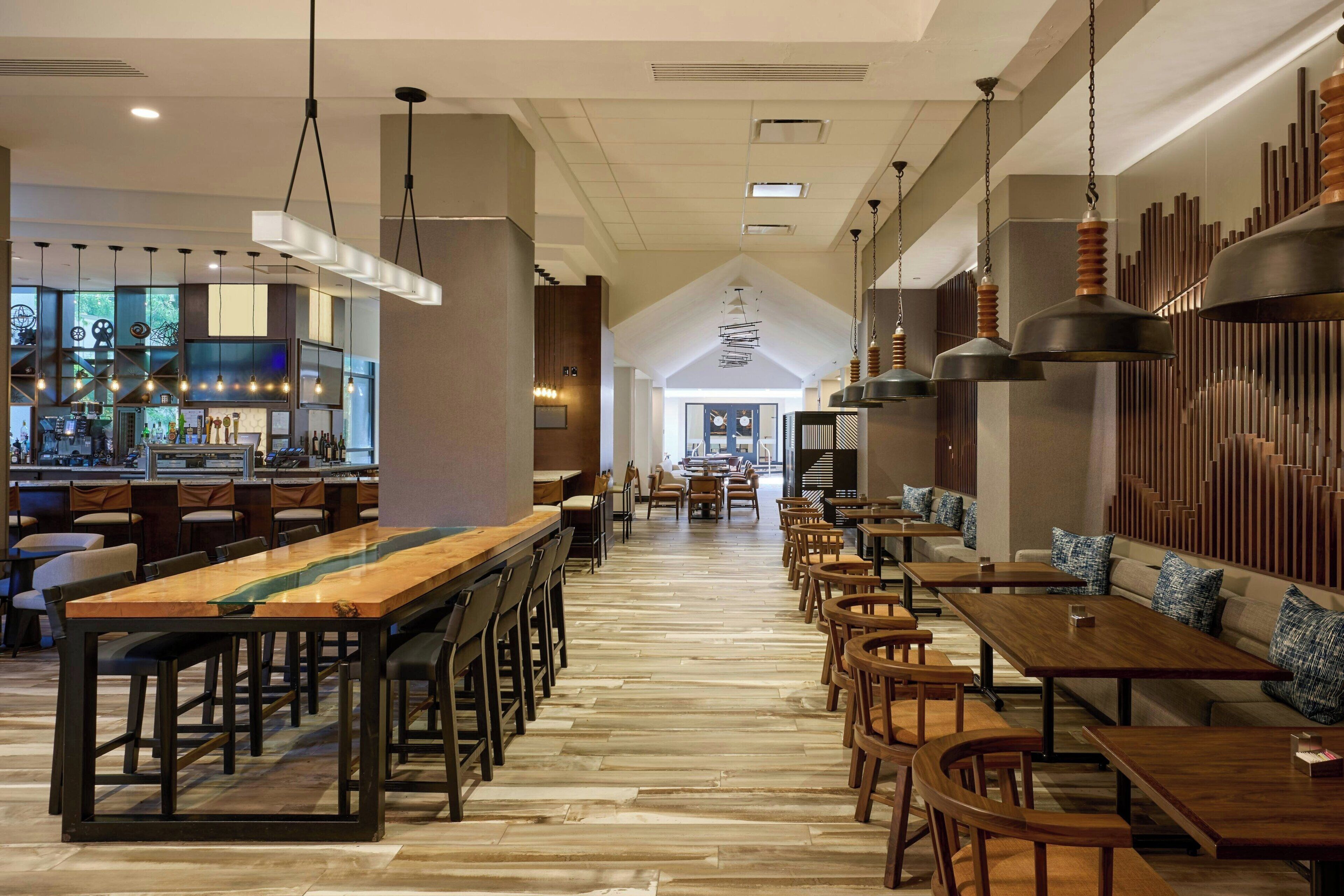 A spacious restaurant with long wooden tables and chairs, a bar area, and a high ceiling with hanging lights.