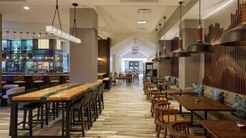 A spacious restaurant with long wooden tables and chairs, a bar area, and a high ceiling with hanging lights.