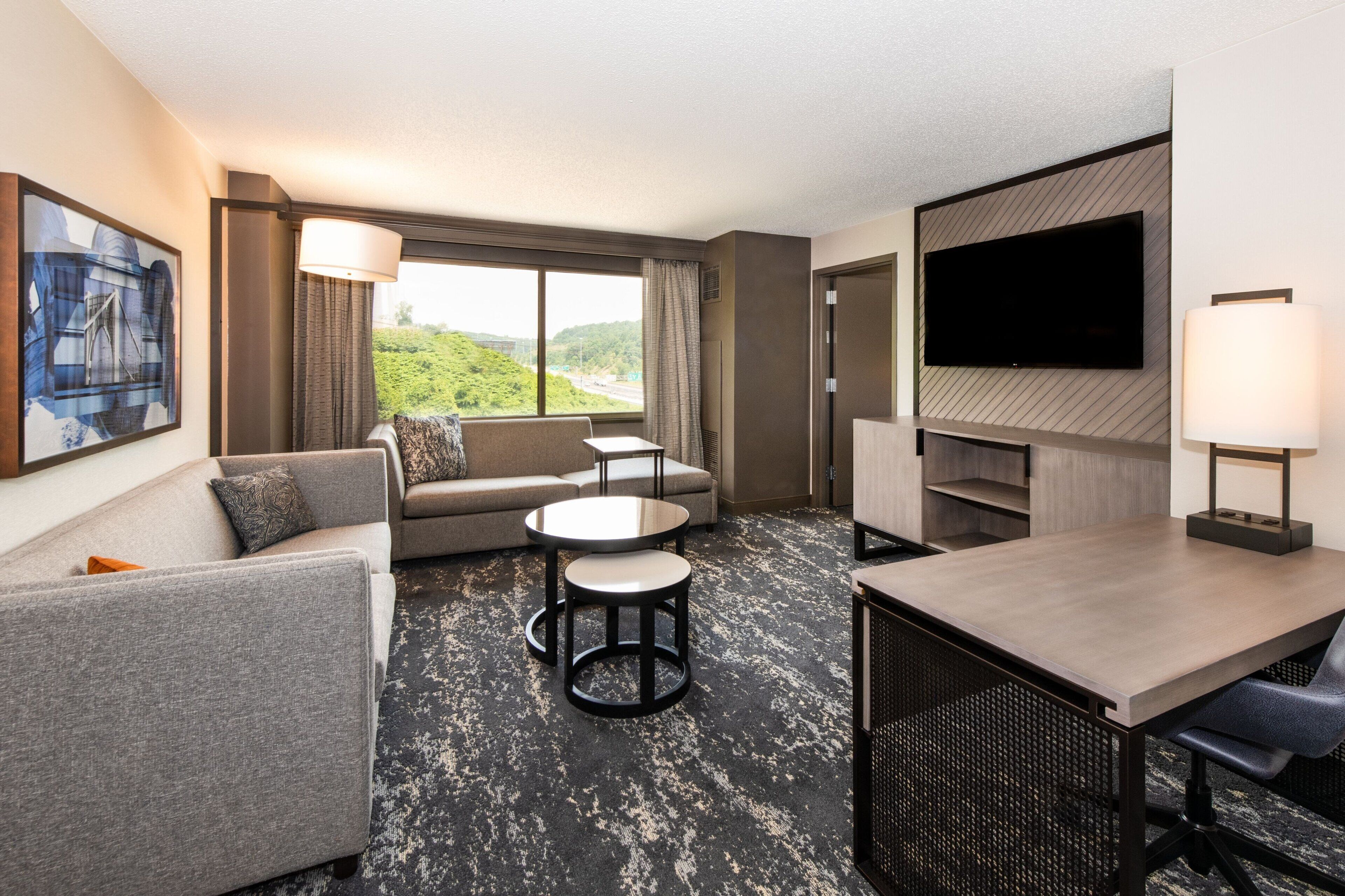 A hotel room with a sofa, two round tables, a TV, and a large window with a view of greenery.