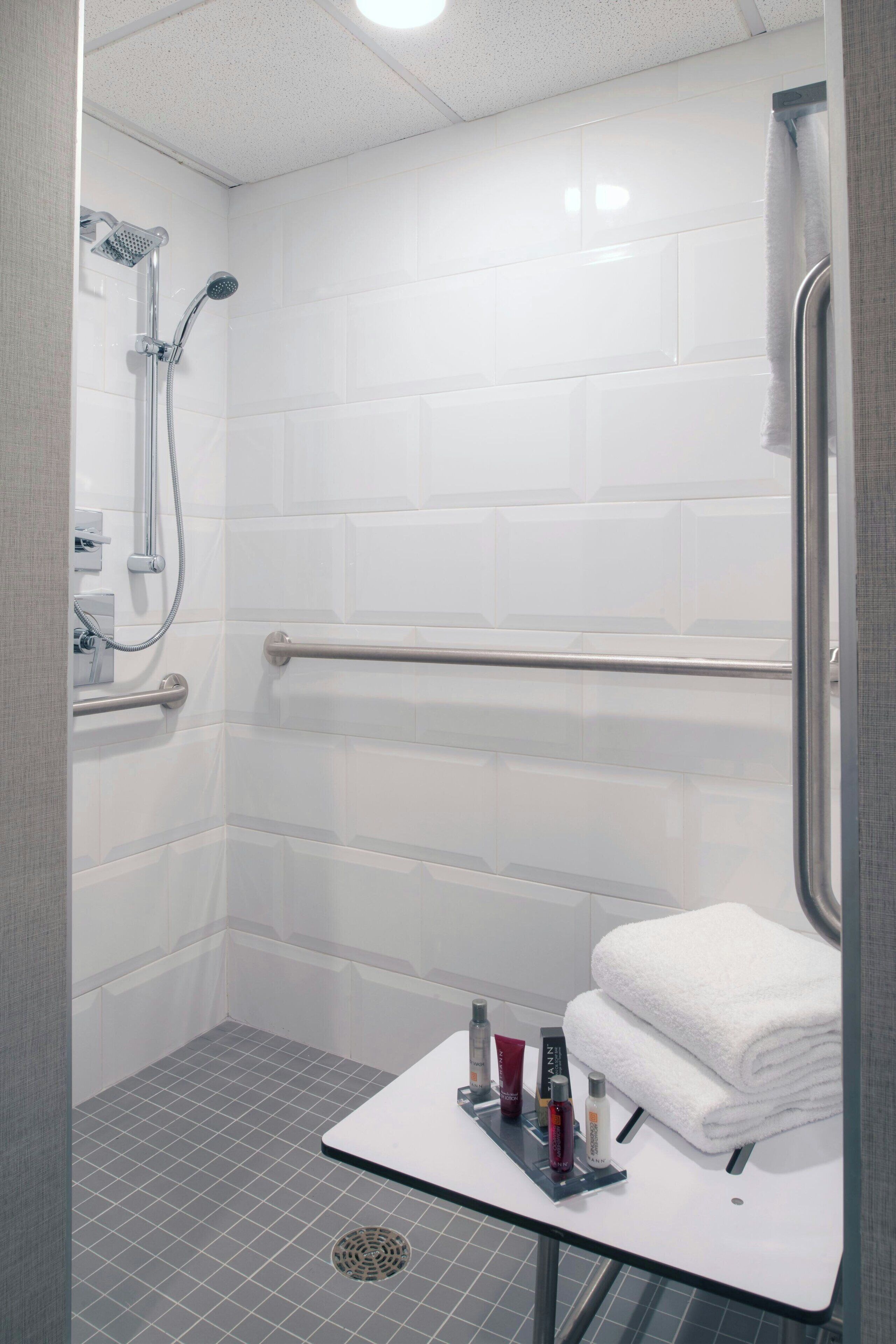 A modern bathroom with white tiles, a shower with a handheld showerhead, a grab bar, and a small table with toiletries.