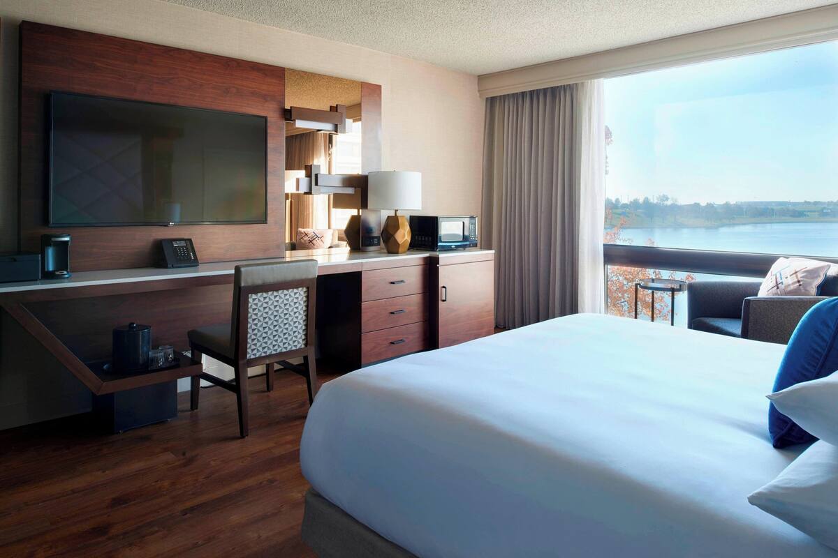 Ein Hotelzimmer mit einem grossen Bett, einem Schreibtisch mit Fernseher, einer Mikrowelle, einer Kaffeemaschine und Blick auf einen See.