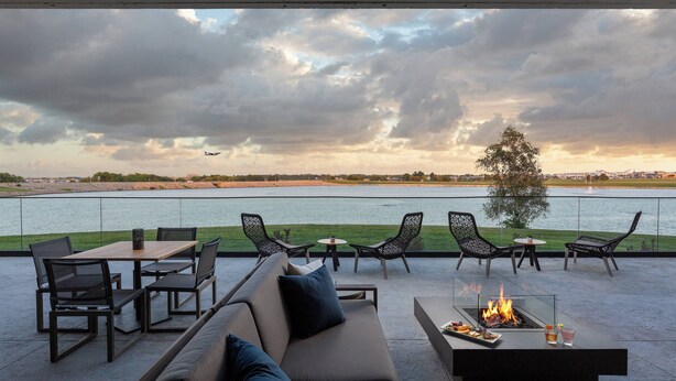 Une terrasse dotée d’un canapé, de chaises et d’une table avec un foyer, offrant une vue sur une étendue d’eau et un paysage lointain.