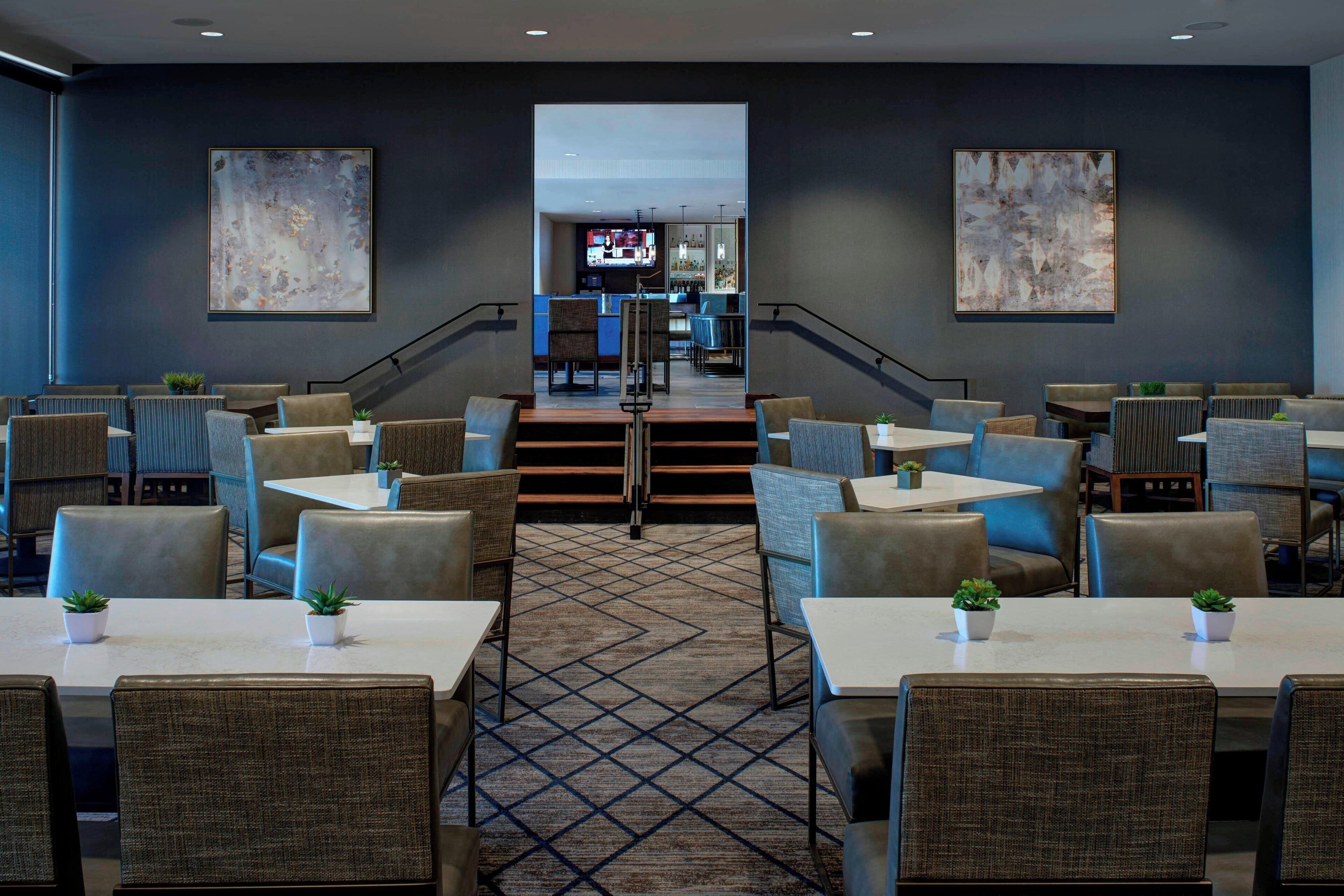 A modern hotel lobby with a central staircase, multiple seating areas, and a bar at the end.