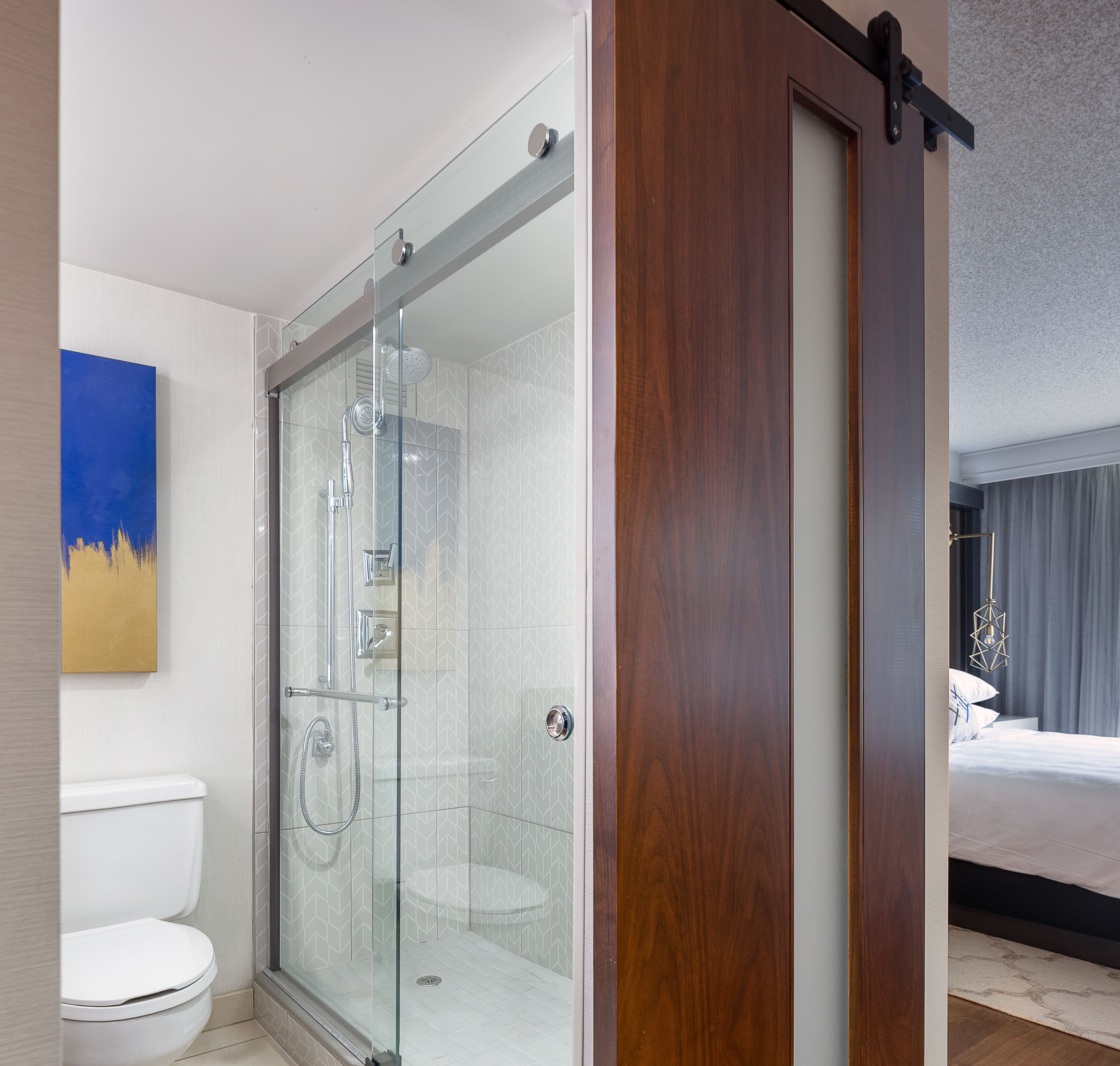 A modern bathroom with a glass-enclosed shower, a toilet, and a bed in the adjacent room.