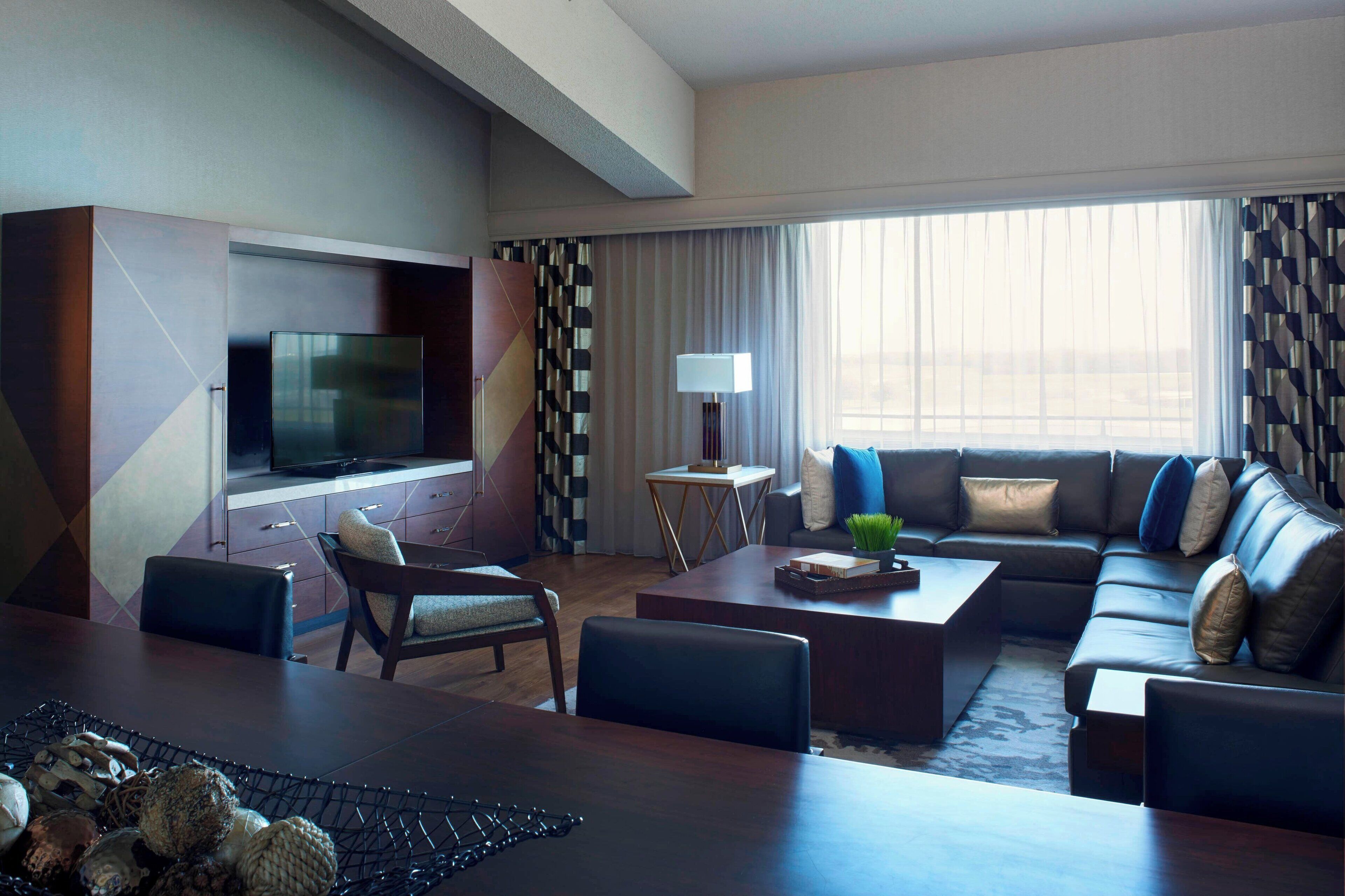 Una habitación de hotel moderna con un televisor de pantalla plana grande, un sofá, una mesa de centro y un comedor con una mesa y sillas de madera.