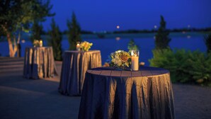 Des tables rondes décorées de bougies et de fleurs, installées à l’extérieur, près d’un point d’eau, au crépuscule.