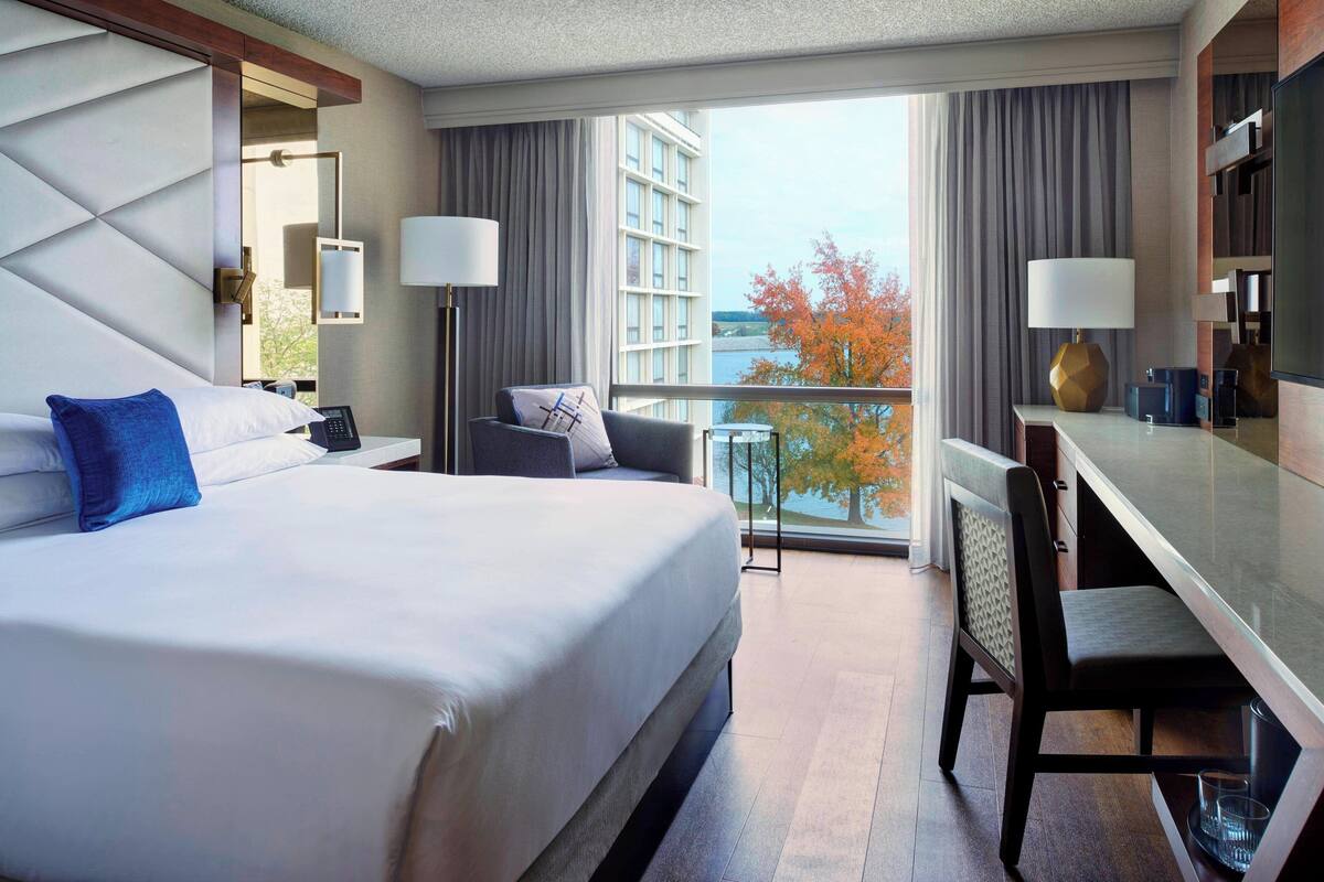 Ein Hotelzimmer mit Bett, Schreibtisch, Stuhl, Fernseher und einem Balkon mit Blick auf die Stadt und Bäume.