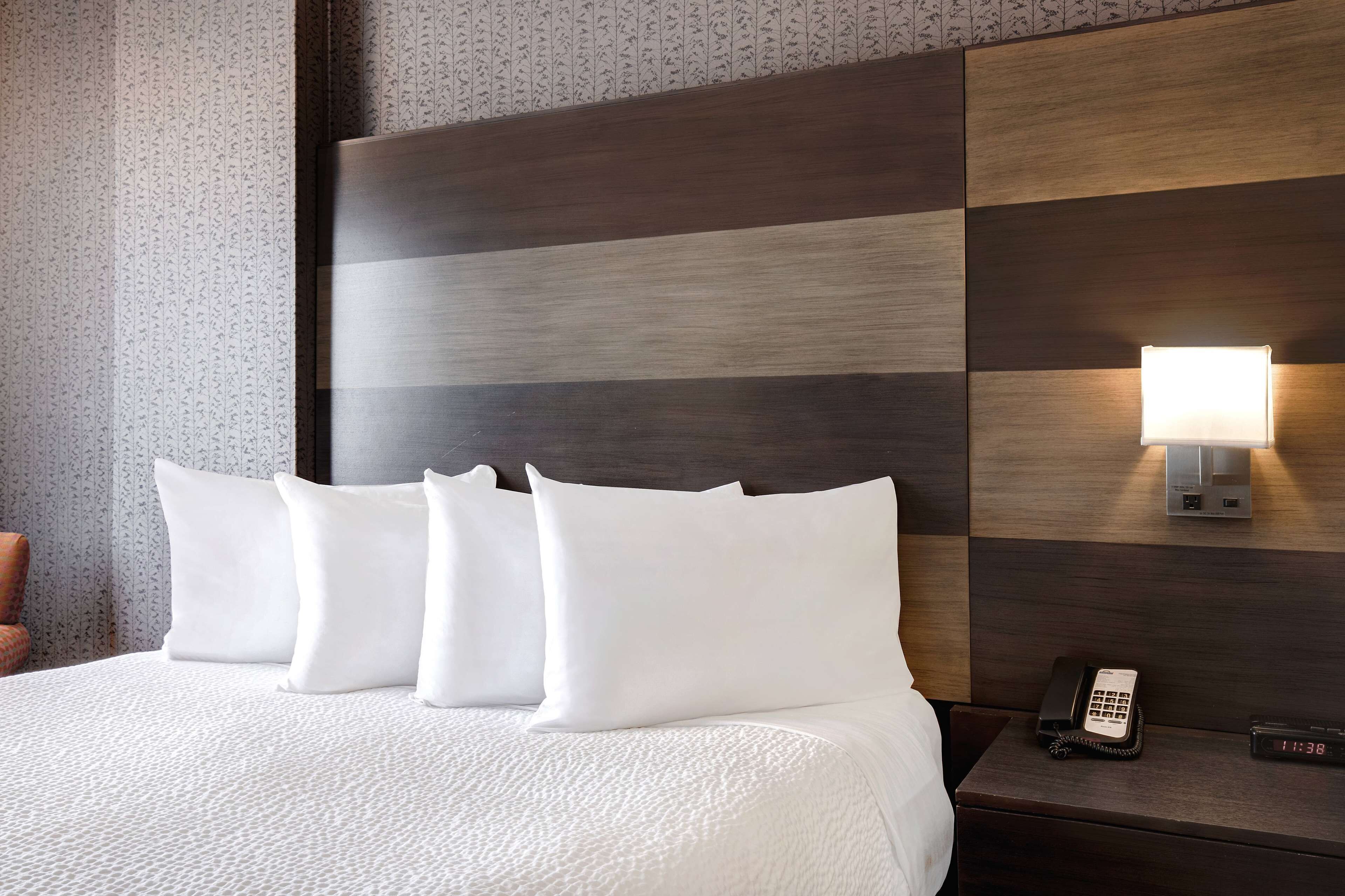 Habitación de hotel con una cama bien hecha, una lámpara de noche, un teléfono y un reloj digital en la mesita.
