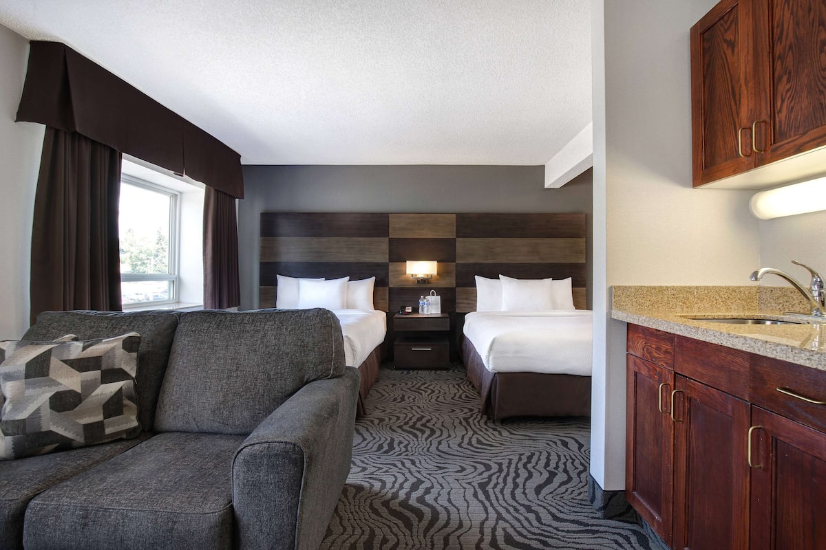 A hotel room with a sofa, two beds, a nightstand with a lamp, a wooden headboard, a kitchen area with a sink, and a window with curtains.