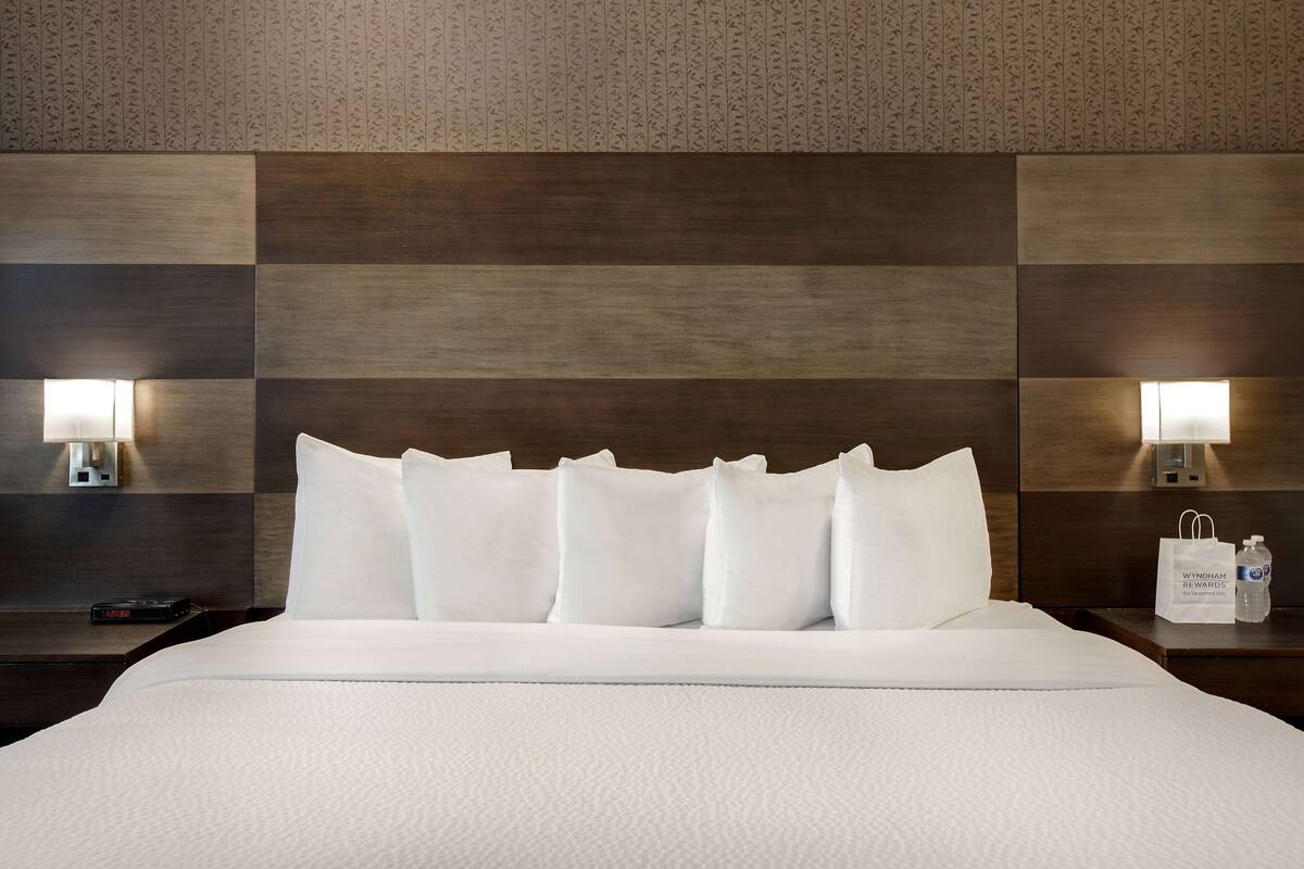 A hotel room with a large bed, white bedding, a wooden headboard, two bedside lamps, and a nightstand with a water bottle and a bag.