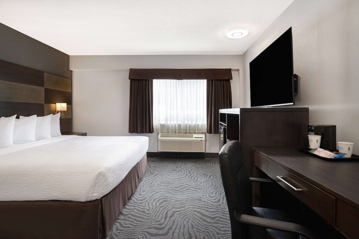 A hotel room with a bed, desk, chair, TV, air conditioning unit, and curtains.