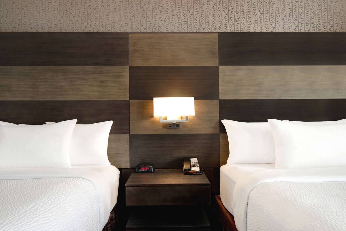 A hotel room with two beds, a nightstand with a clock and phone, and a wall-mounted light fixture.