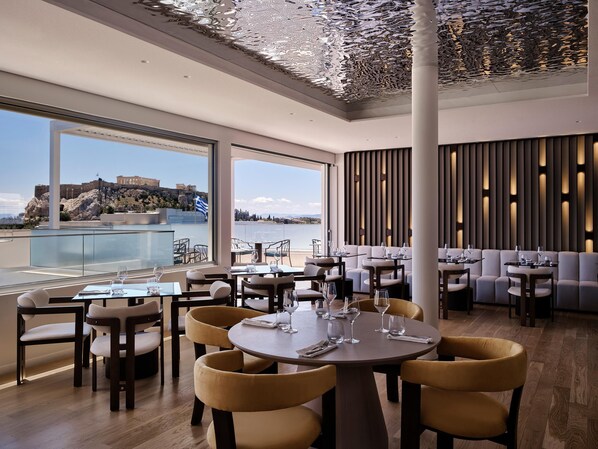 A modern restaurant with round tables and yellow chairs, large windows offering a view of a historic building and the sea.