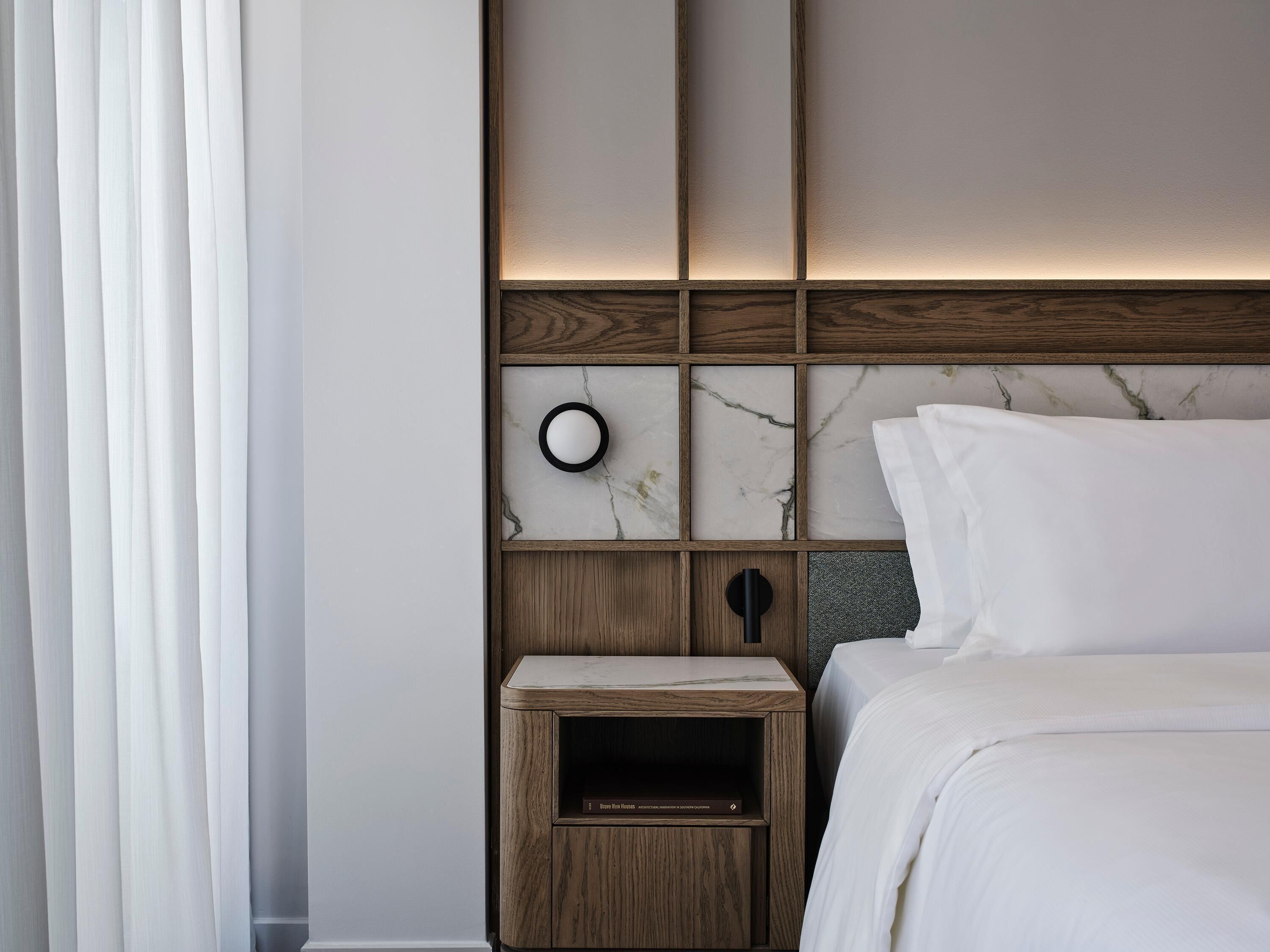 Una habitación de hotel con cabecera de madera, pared con diseño de mármol, mesita de noche y cama con ropa blanca.