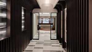 A modern hallway with a sleek, dark interior design, featuring a glass partition leading to a well-lit room with a seating area.
