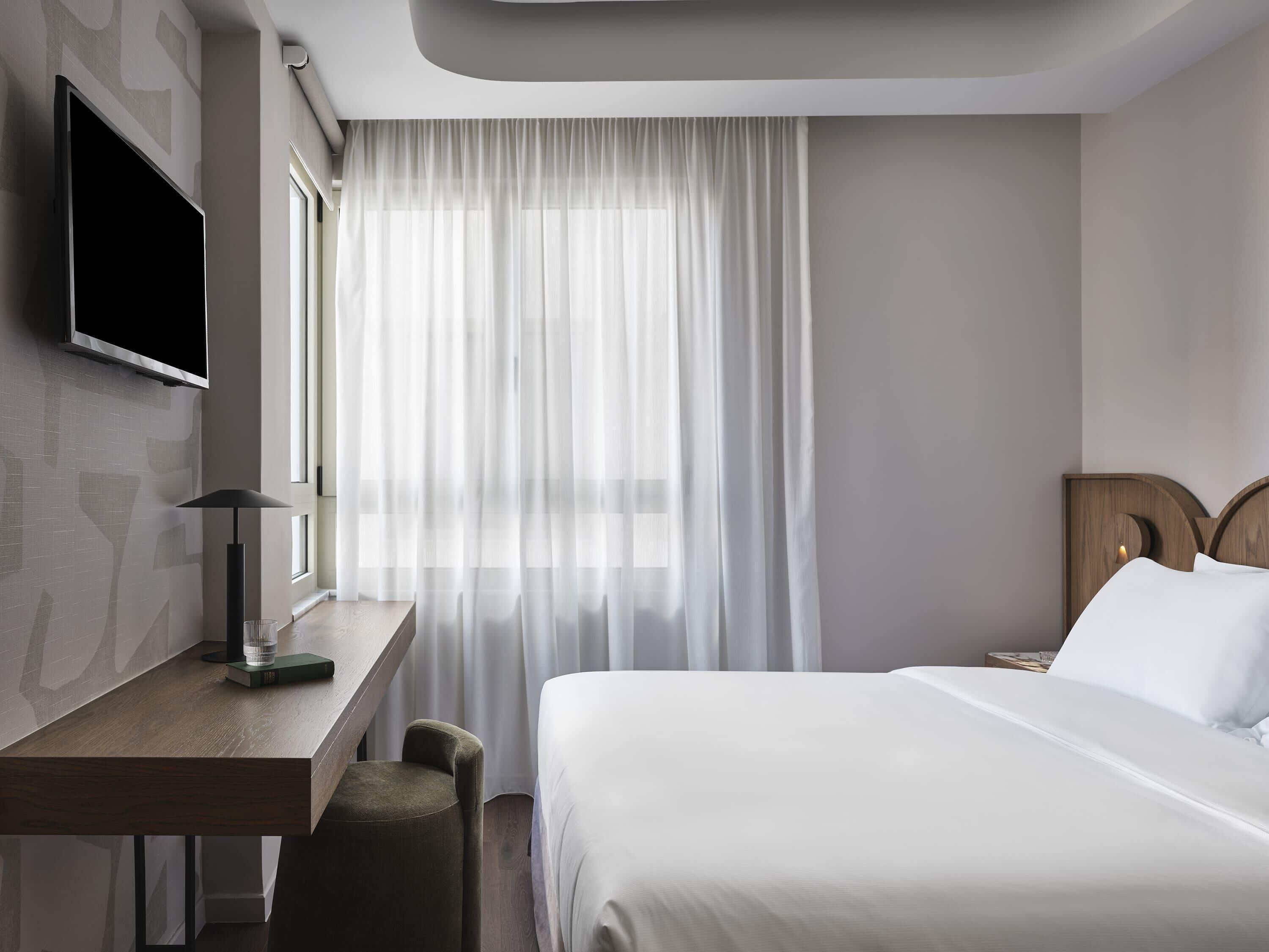Una habitación de hotel con cama, escritorio, televisor montado en la pared y ventana con cortinas translúcidas.