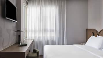 Una habitación de hotel con cama, escritorio, televisor montado en la pared y ventana con cortinas translúcidas.