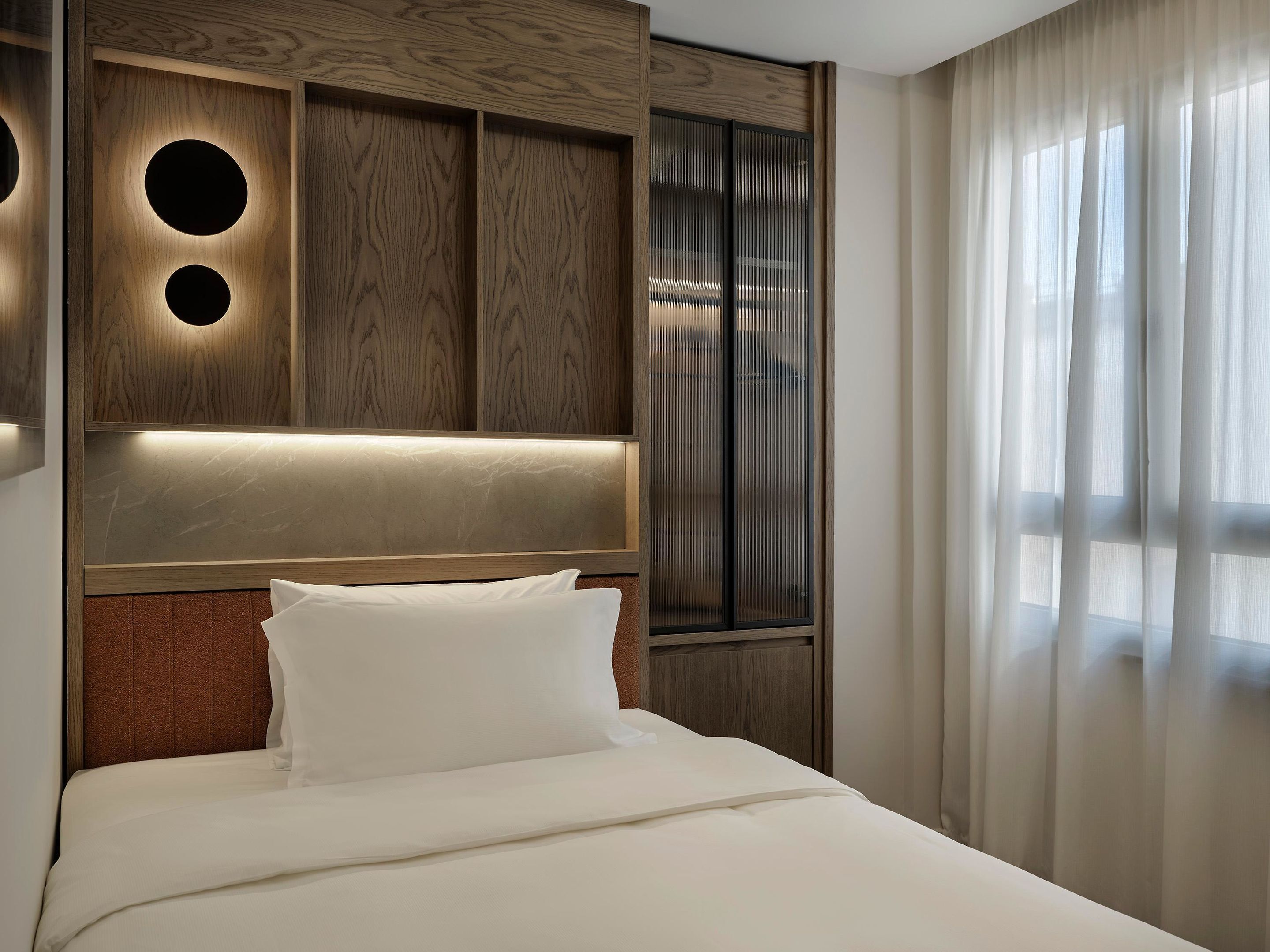 A hotel room with a bed, a headboard with built-in lighting, a large window with sheer curtains, and a wooden panel wall design.