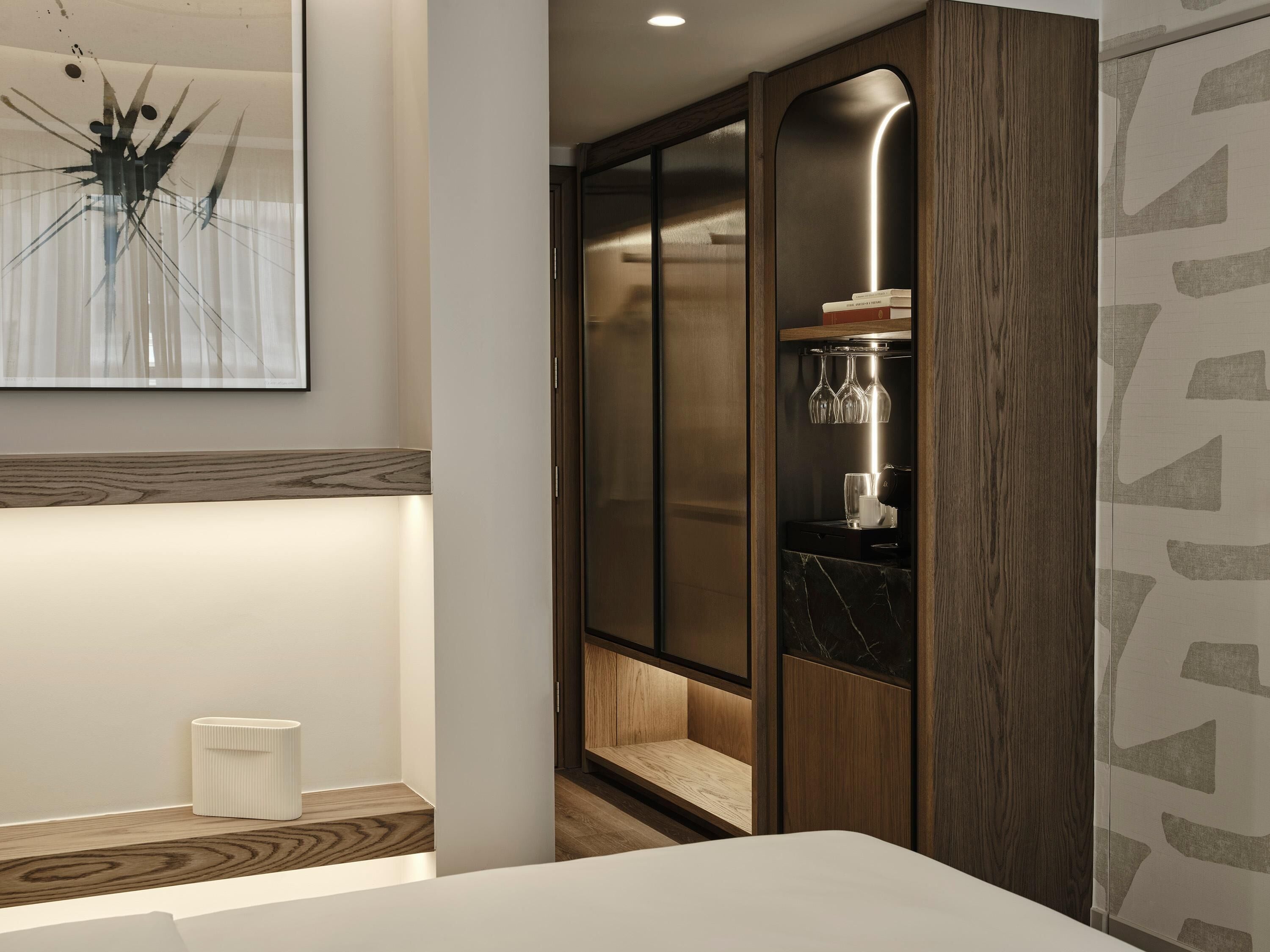 A modern hotel room with a wooden vanity, a large framed artwork, and a built-in wardrobe with a wine glass holder.