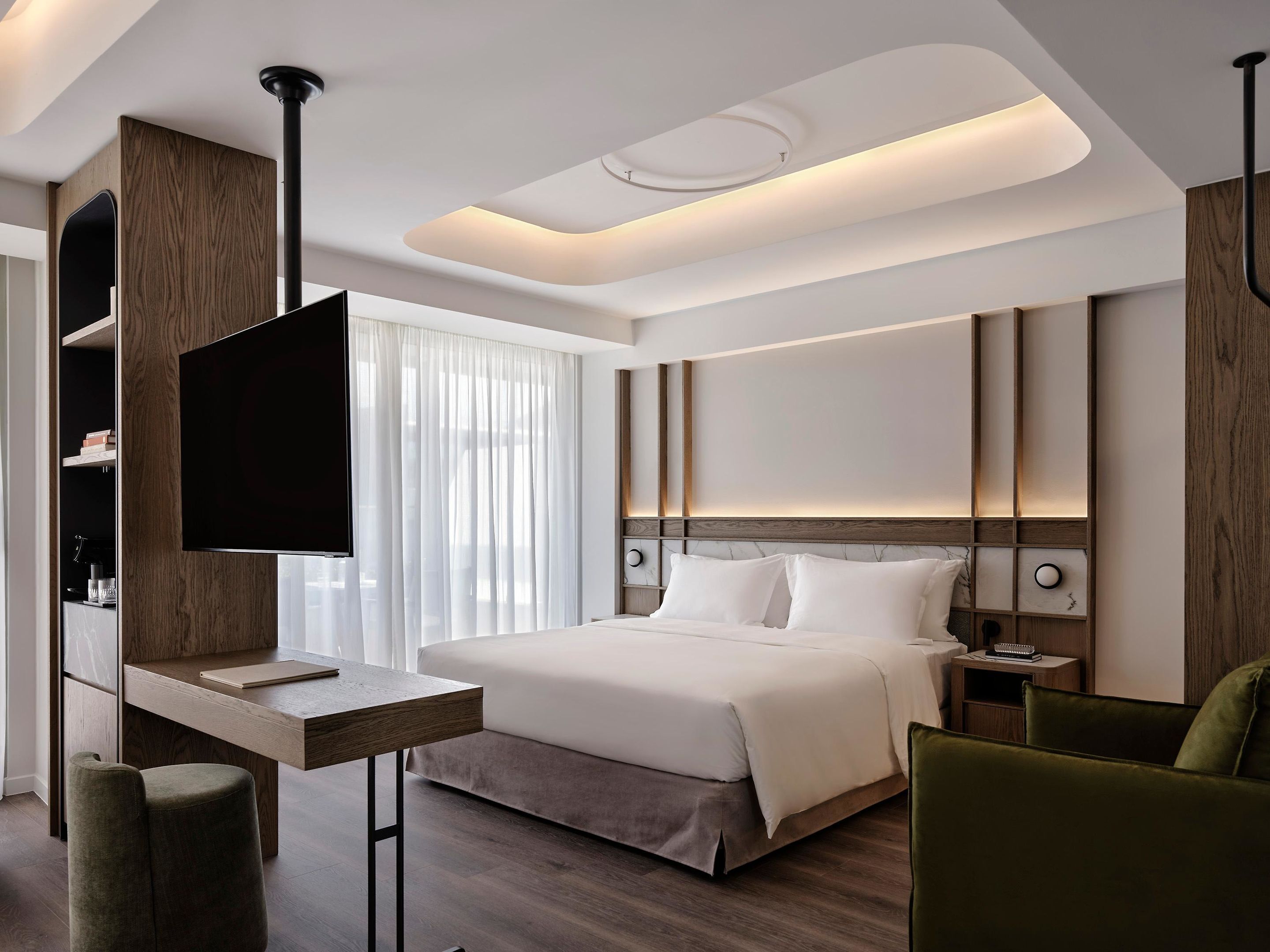Quarto de hotel moderno com uma cama grande, televisão de tela plana fixada em um painel de madeira, mesa de cabeceira e uma poltrona verde.