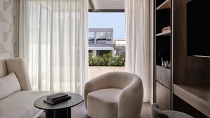 A modern living room with a sofa, armchair, and a small table with books. A large window with sheer curtains allows natural light and a view of a building outside.