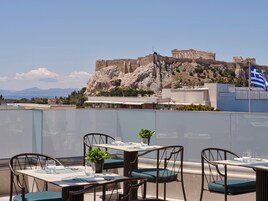 Terraza en la azotea con mesas y sillas, con vistas a ruinas antiguas y un edificio moderno con una bandera.