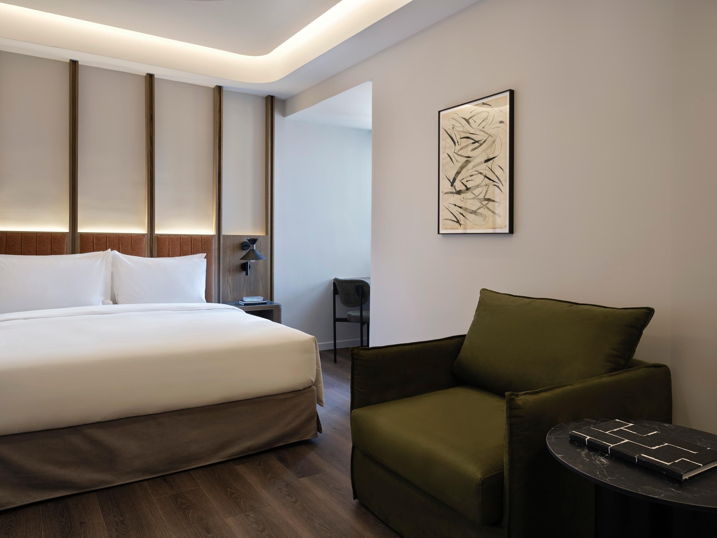 A modern hotel room with a large bed, a green armchair, a small table, and a wall-mounted abstract art piece.
