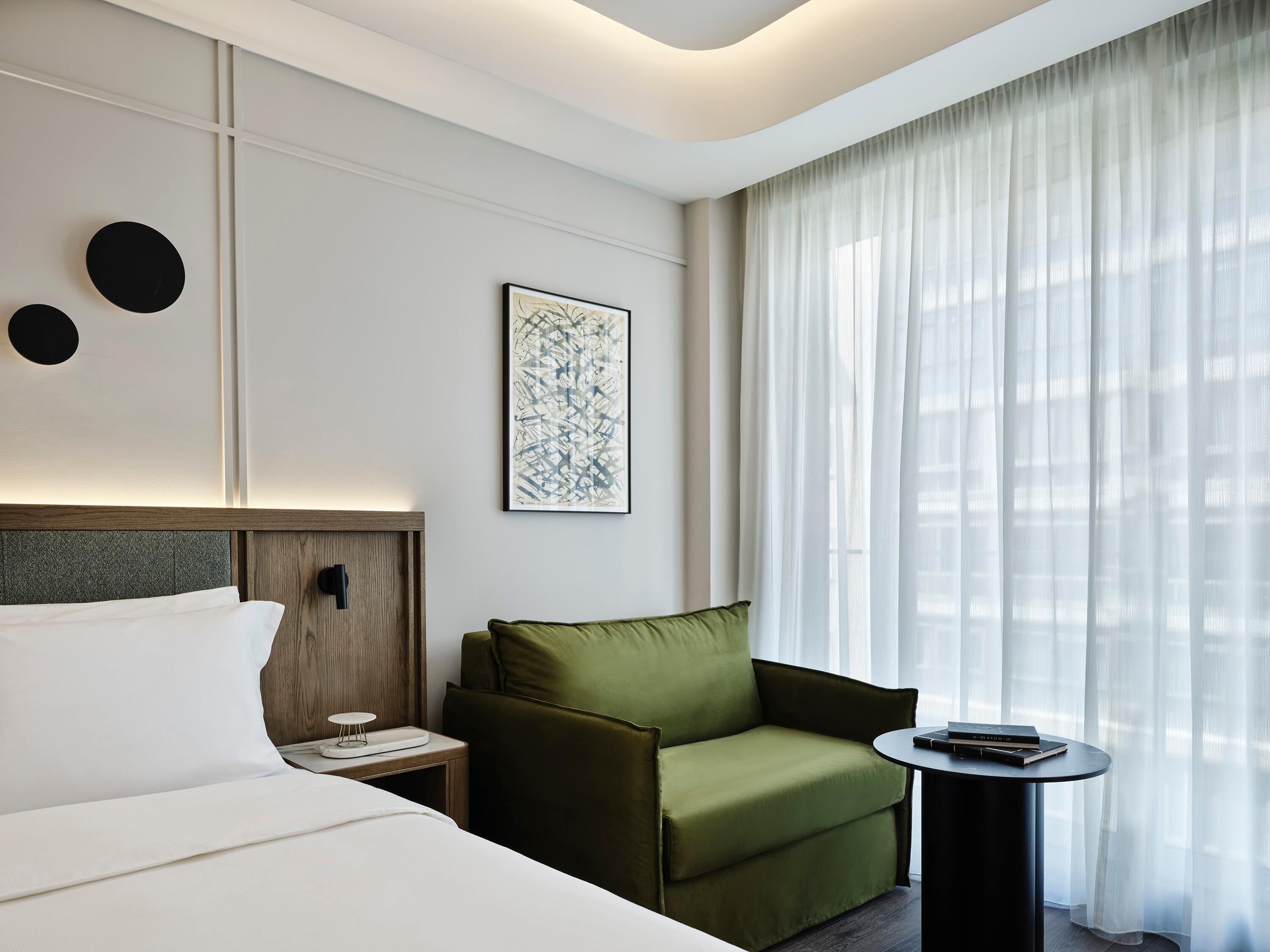 A modern hotel room with a bed, a green armchair, a small table, and a large window with sheer curtains.