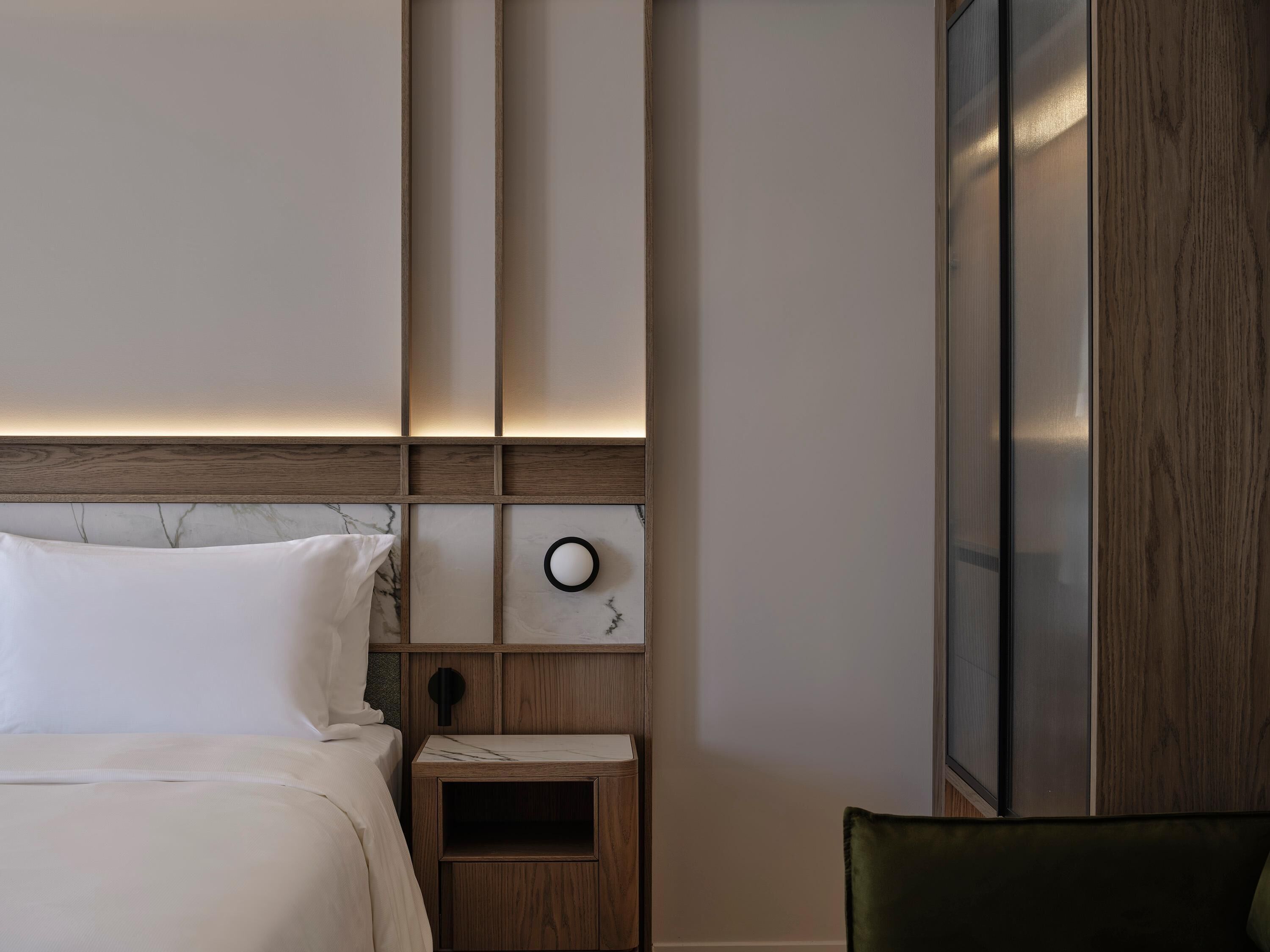 Una habitación de hotel con cama, mesita de noche, un reloj en la pared y detalles de panel de madera.