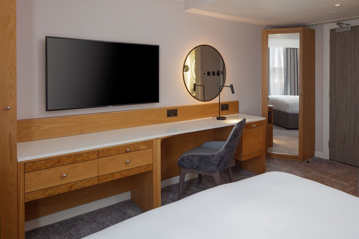 A modern hotel room with a large bed, a wooden desk, a flat-screen TV, a round mirror, and a walk-in closet.